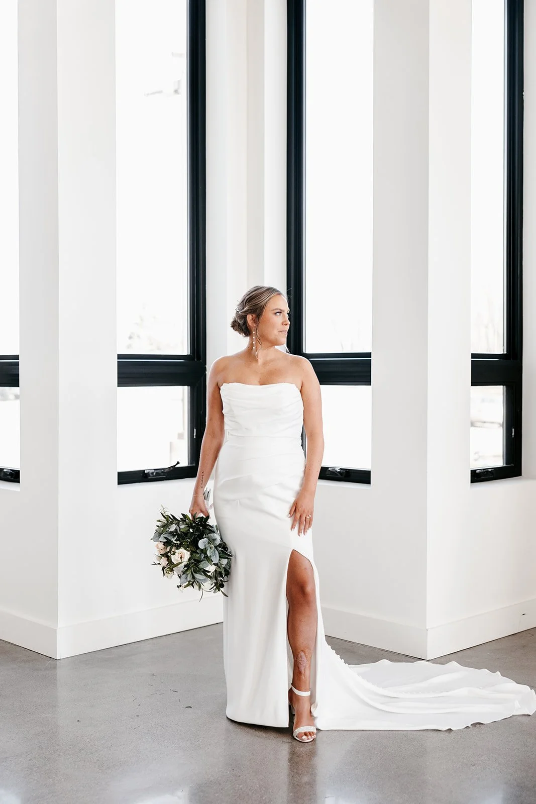 A bride in a strapless white wedding gown holding a bouquet of flowers, standing indoors near large black window frames, looking to her left.