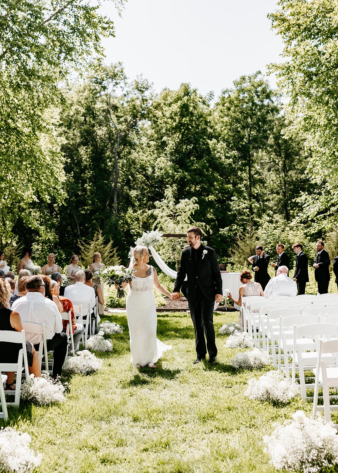 A couple gets married outdoors on a grassy area surrounded by trees, walking hand in hand down the aisle with guests seated on either side, flowers decorate the aisle, and a wedding party stands in the background.