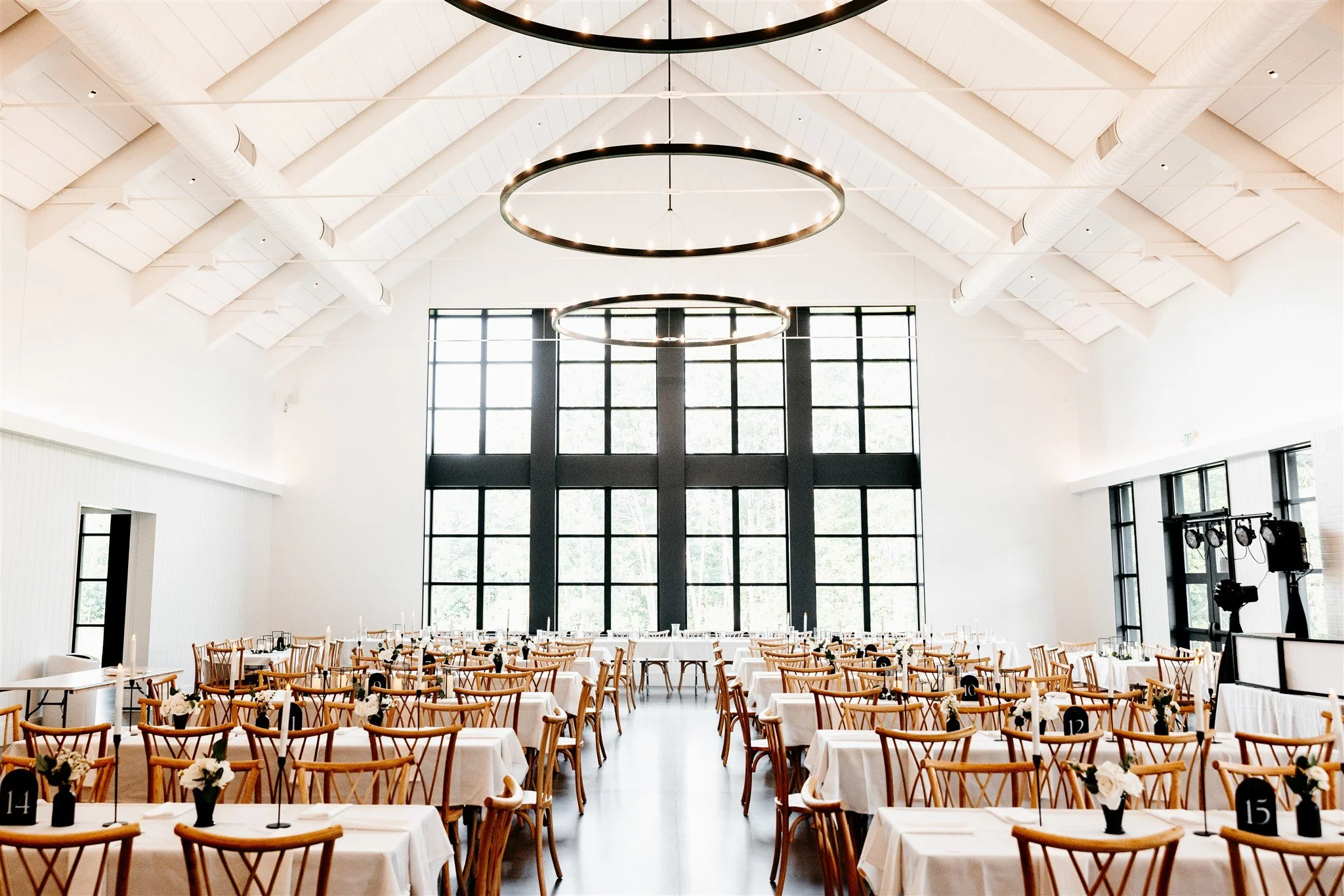 A spacious, modern event hall with high vaulted ceilings and large windows. The hall is set up for a formal event with multiple tables covered in white tablecloths, decorated with small flower arrangements in black vases. Wooden chairs are arranged a