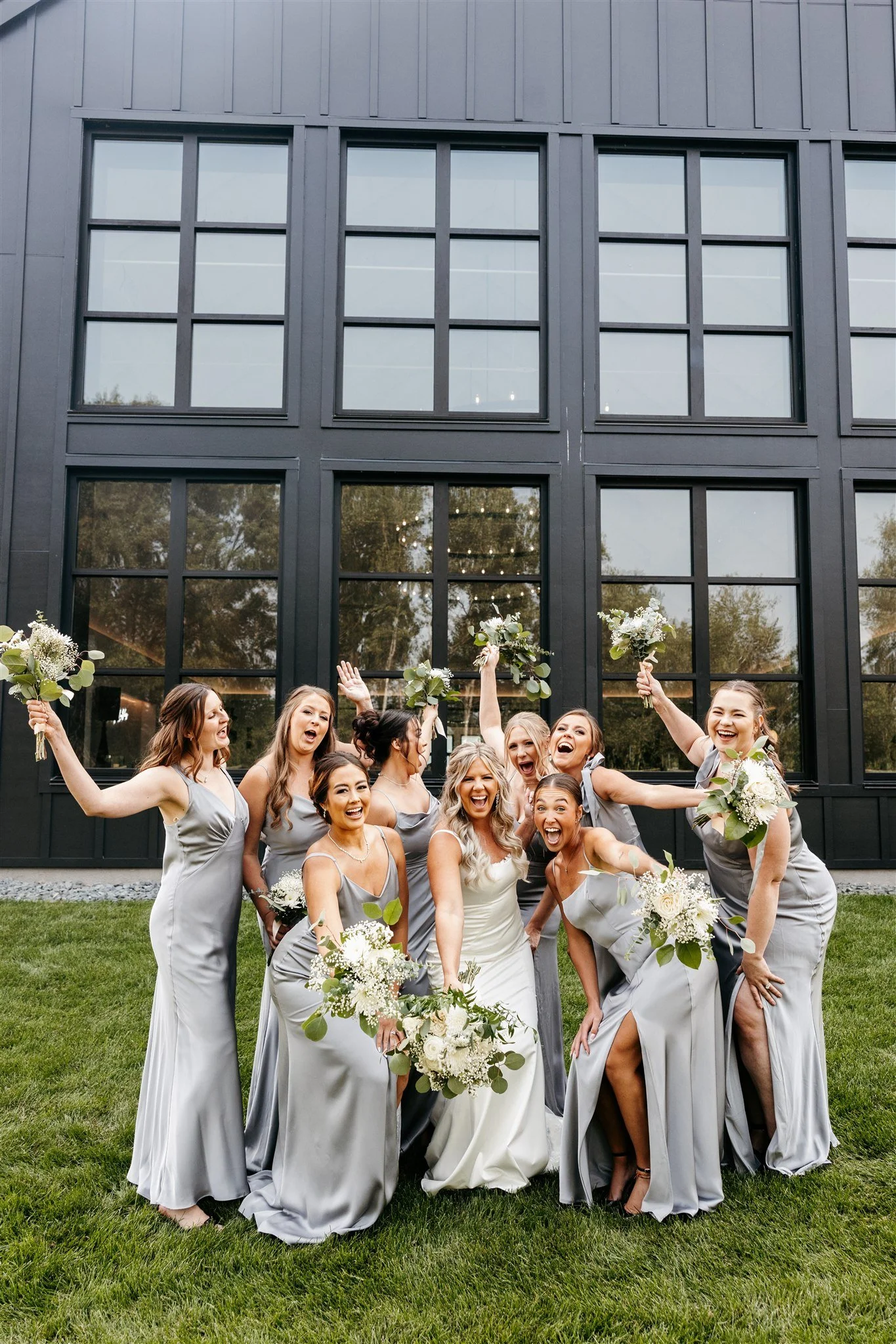 A group of women dressed in matching gray dresses, holding bouquets of white flowers, celebrating and smiling outdoors in front of a black building with large windows.