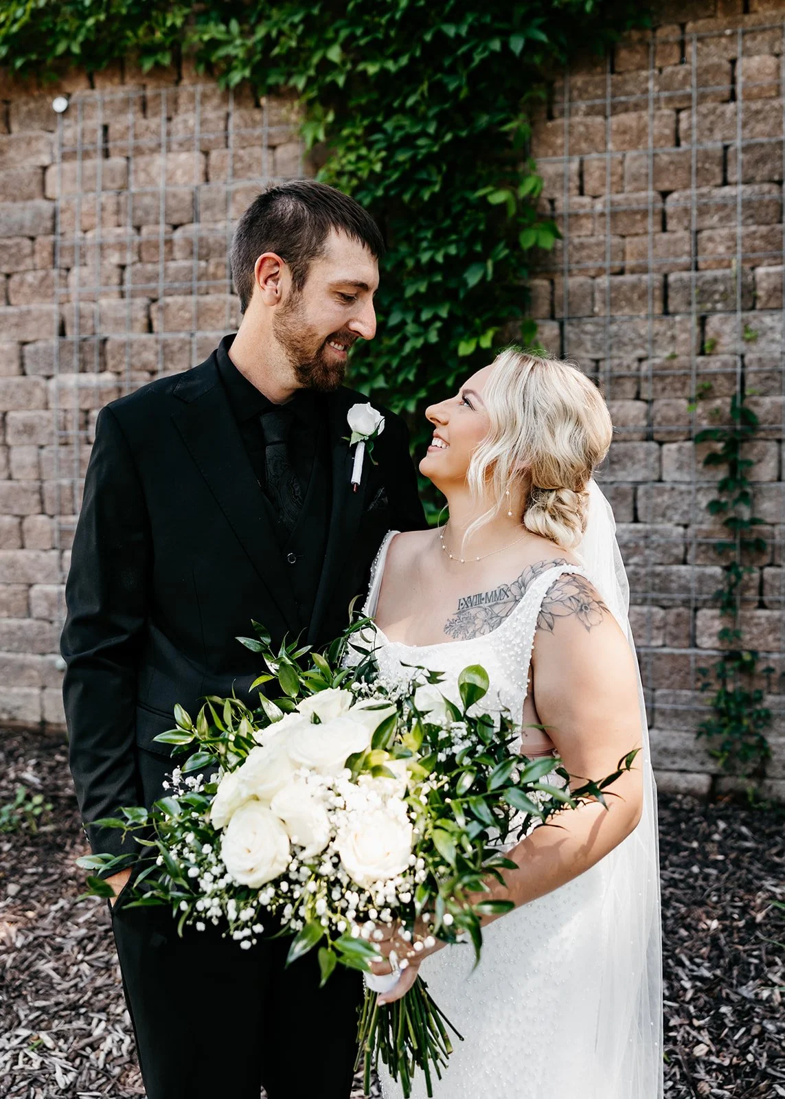 A bride and groom smiling at each other outdoors, with the bride holding a bouquet of white roses and greenery, against a brick wall with green vines.