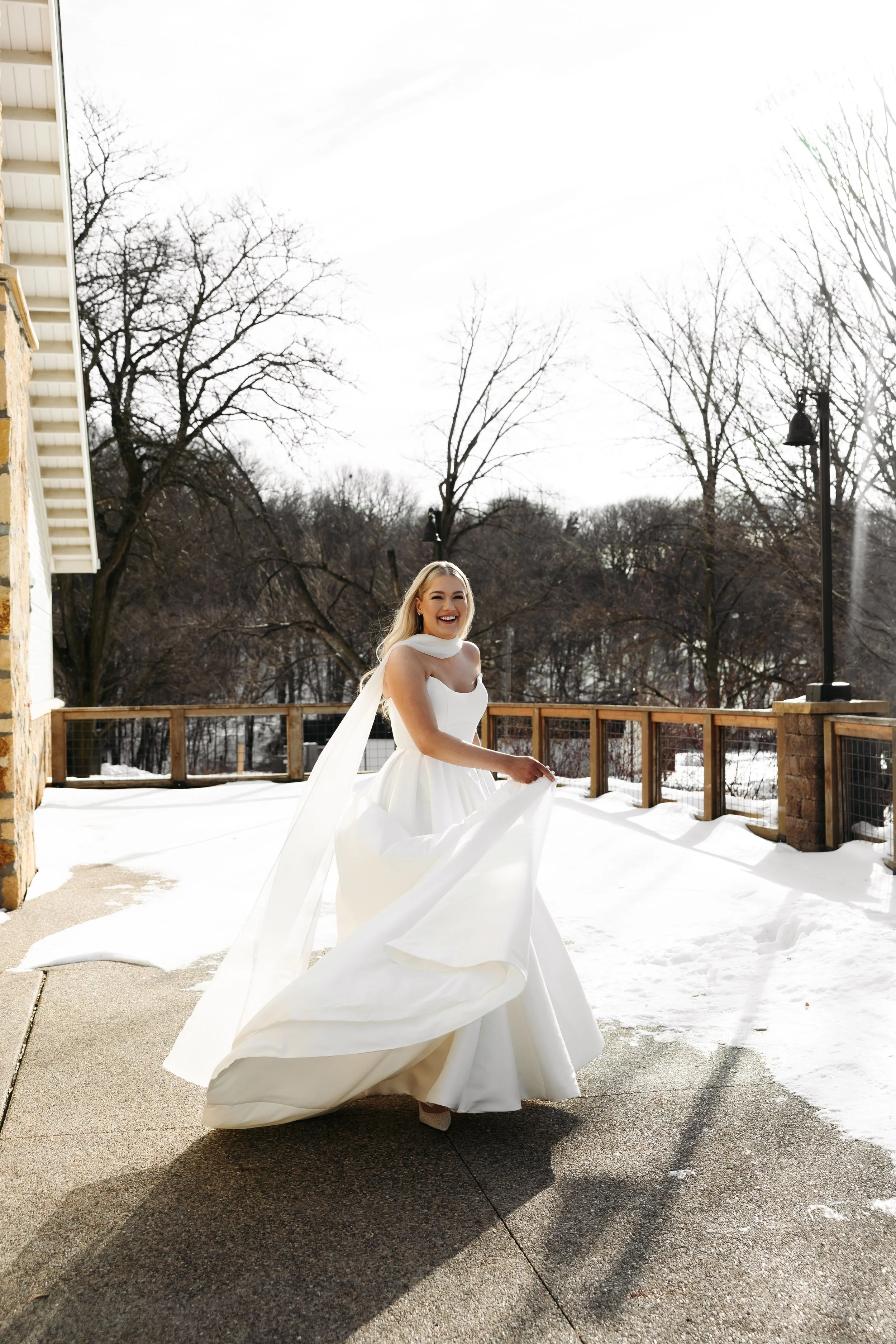 A woman in a wedding dress standing on a snow-covered outdoor patio, smiling and holding up her dress.