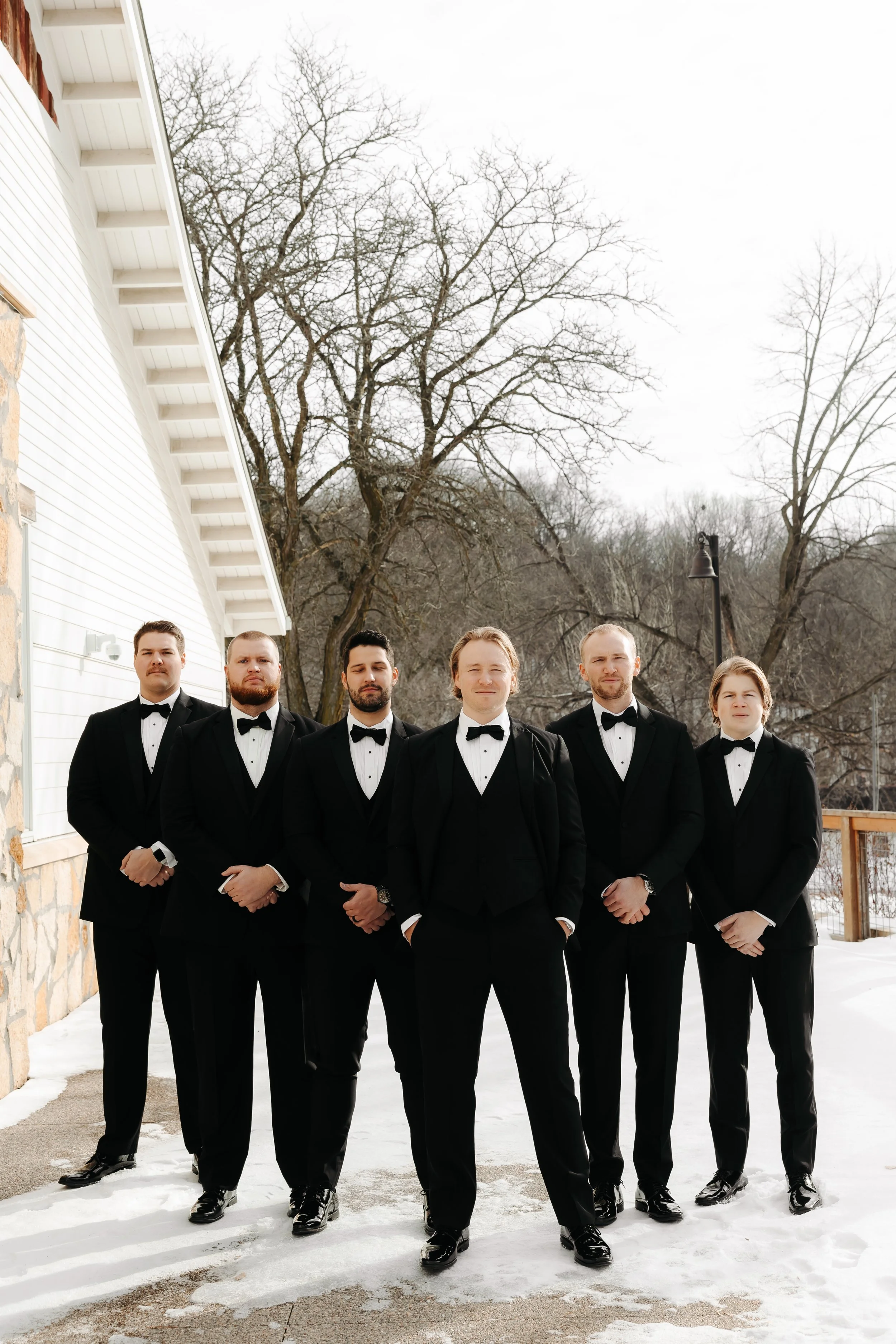 Group of seven men in tuxedos standing outdoors on snow-covered ground with leafless trees in the background.