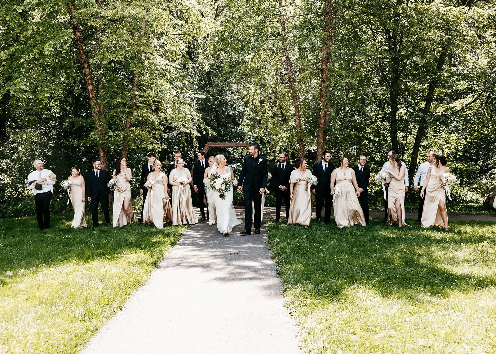 Bride and groom walking hand-in-hand in front of wedding party on a gravel path surrounded by green trees.