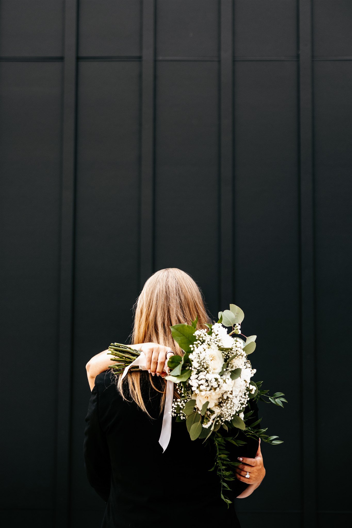 A woman with long blonde hair dressed in black, holding a large bouquet of white flowers and greenery over her shoulder, standing against a black wall.