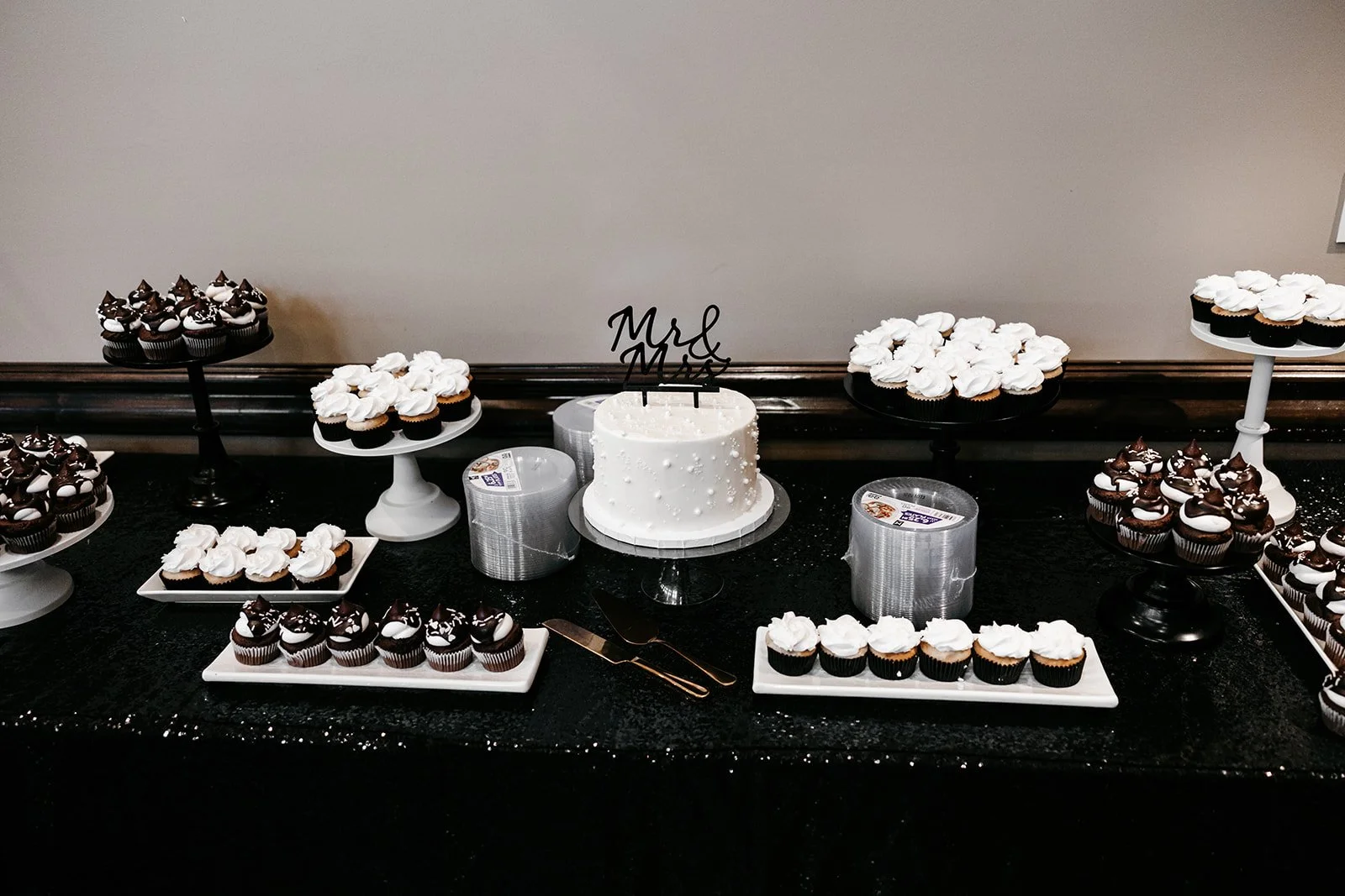 Wedding dessert table with white and chocolate cupcakes, a white wedding cake with 'Mr & Mrs' topper, set on black and white stands with some empty cake containers below.