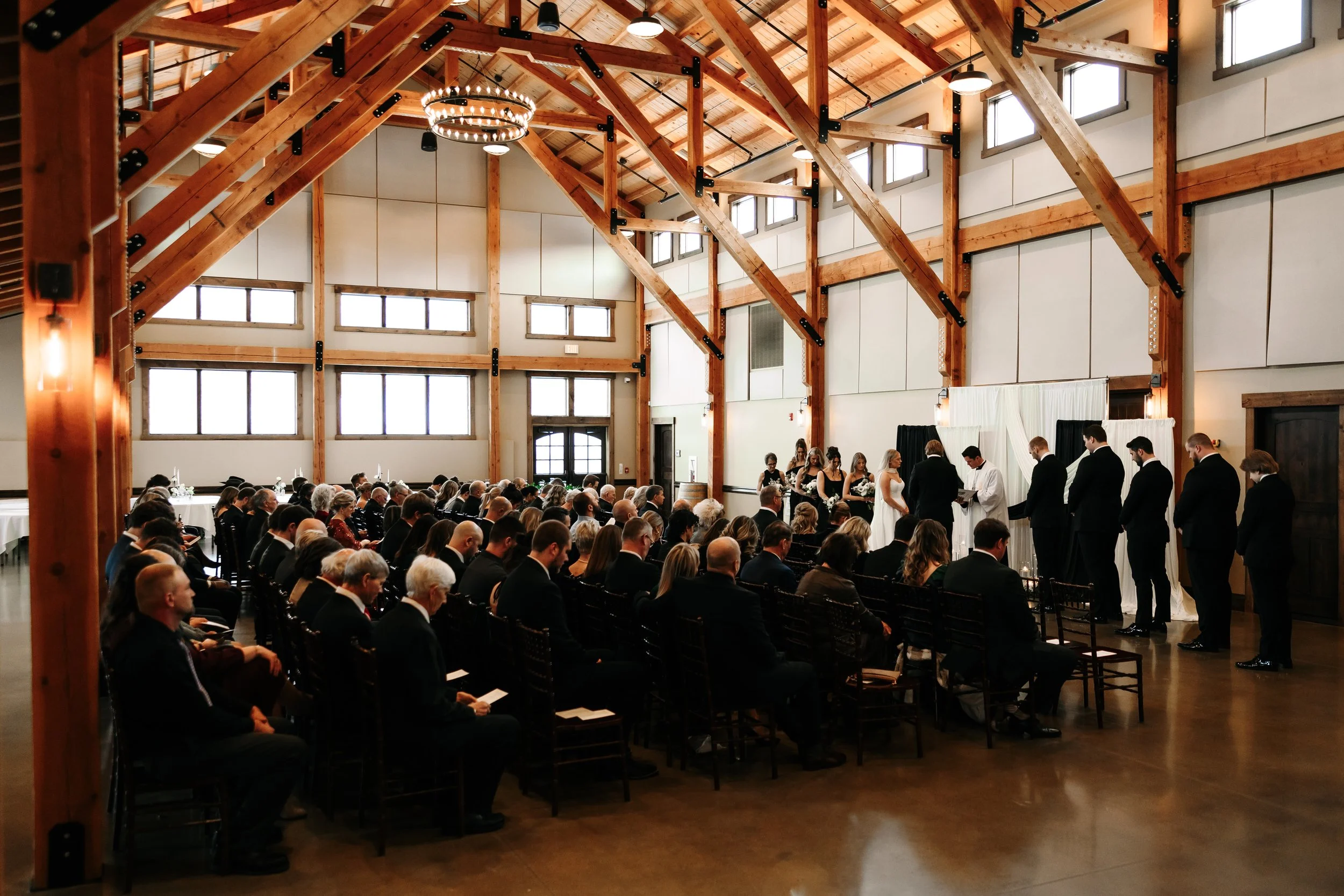 A wedding ceremony inside a wooden barn with high ceilings, where guests are seated and the bride and groom are standing at the altar with a group of bridesmaids and groomsmen.