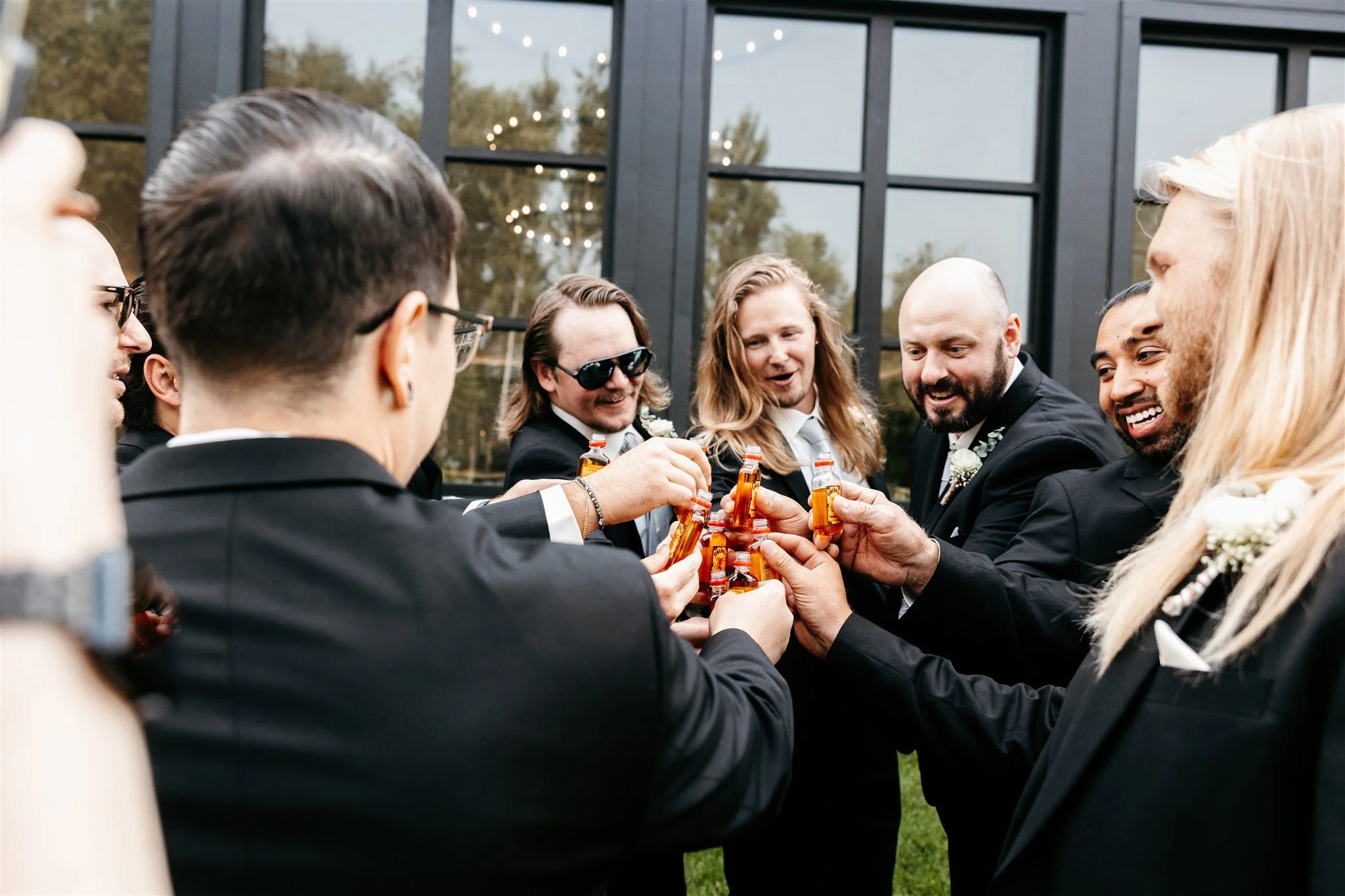 A group of men dressed in tuxedos clinking bottles of beer together in an outdoor setting.