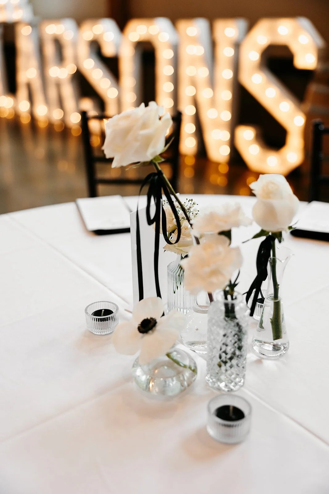 Elegant table centerpiece with white flowers in clear glass vases, set on a white tablecloth, with blurred warm glowing marquee lights spelling out 'LOVE' in the background.