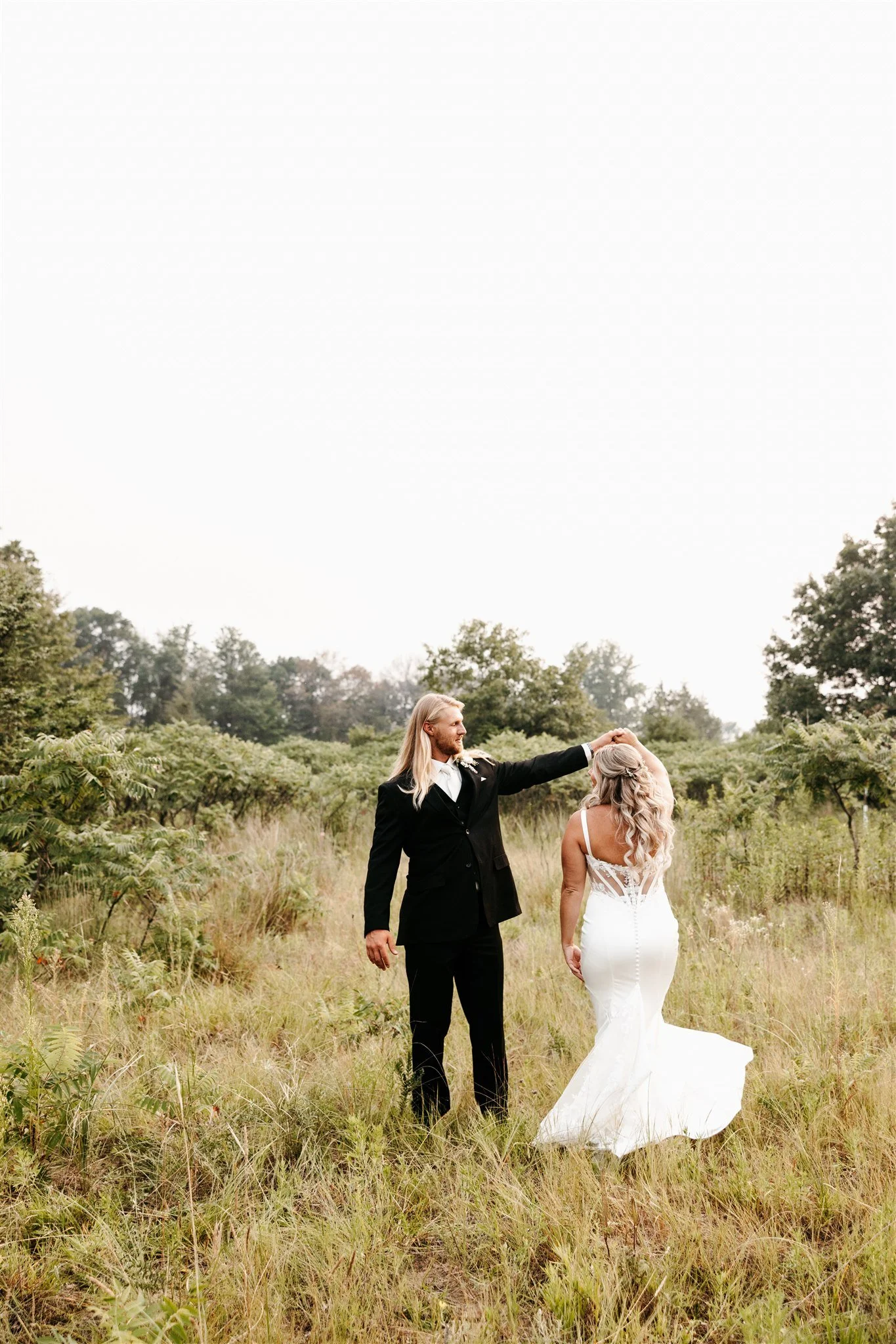 A bride and groom dancing in a field with green trees, outdoor and overcast sky background.