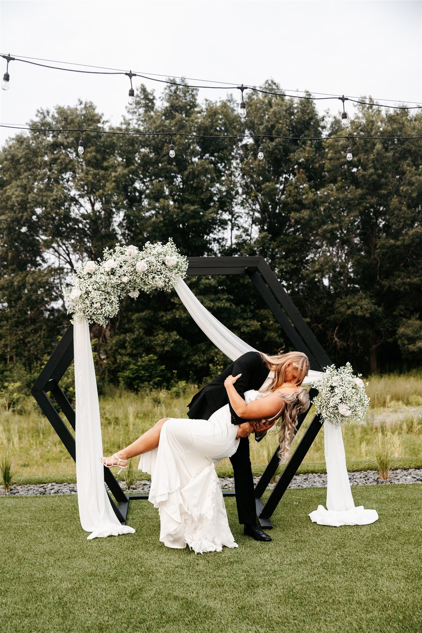 A bride and groom share a kiss during their outdoor wedding ceremony with a modern geometric arch decorated with white flowers and drapery, set against a grassy field and trees.