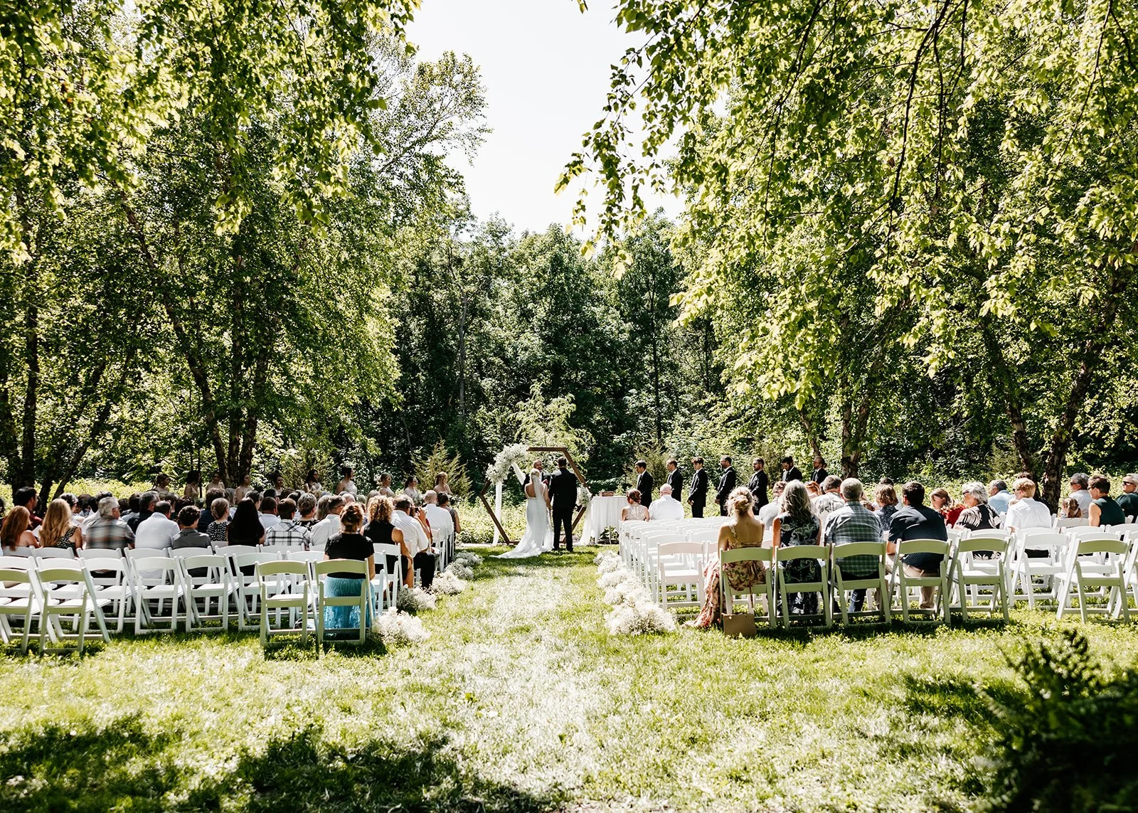 Outdoor wedding ceremony under trees with guests seated in white chairs, bride and groom at the altar.
