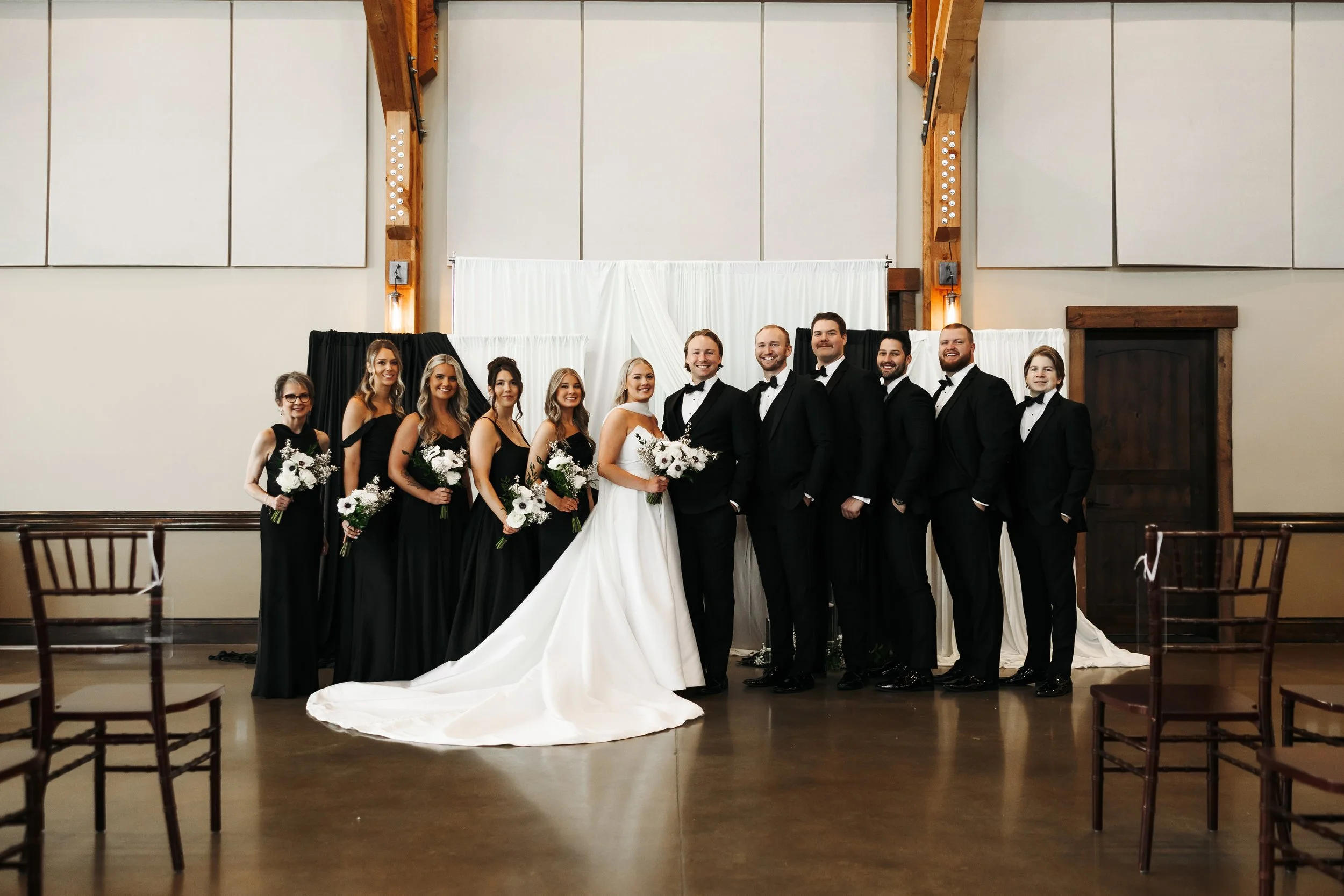 A wedding party with the bride and groom in the center, dressed in a white wedding gown and black tuxedo, respectively, surrounded by bridesmaids in black dresses and groomsmen in black tuxedos, all holding bouquets of white flowers, standing inside 