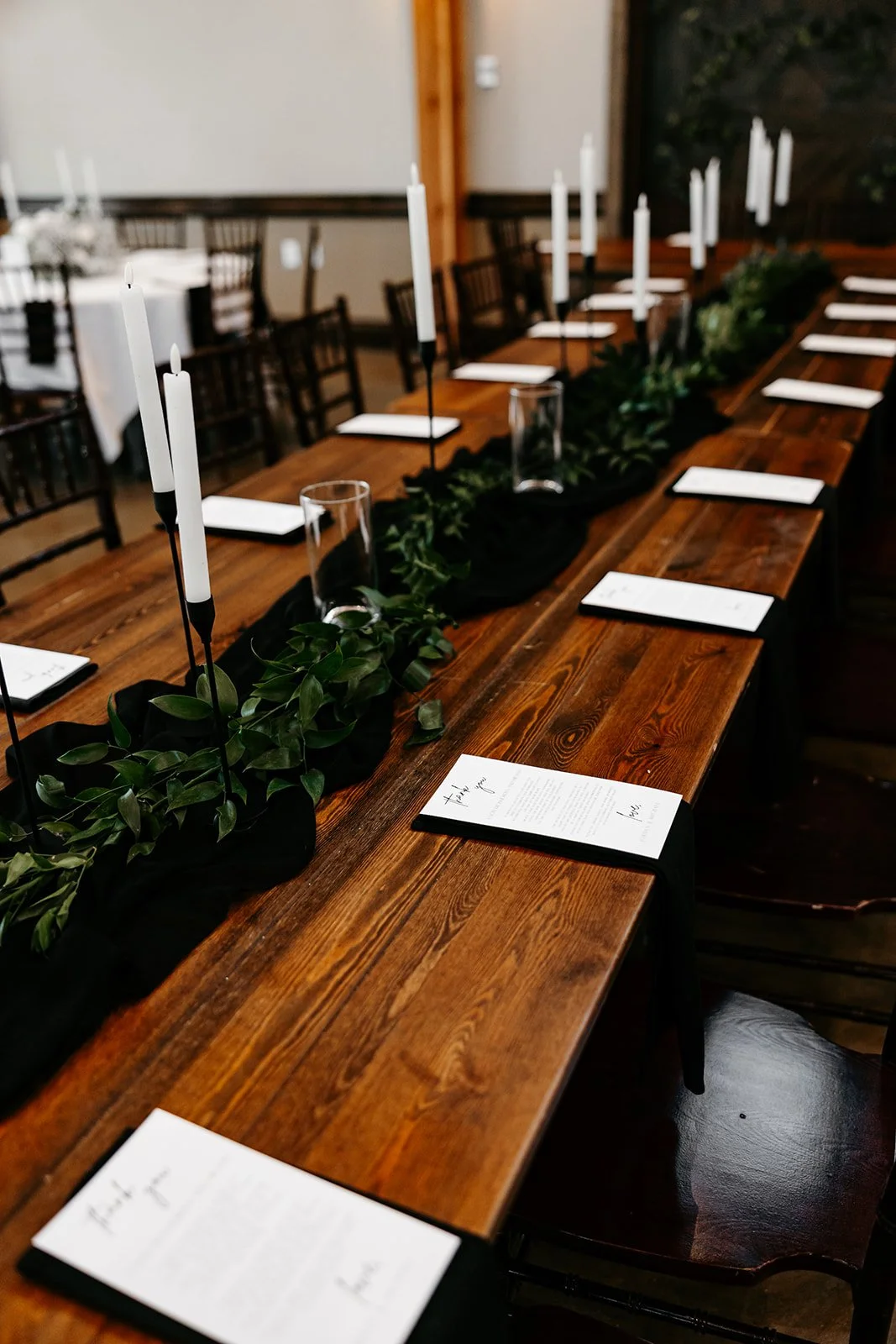 A long wooden table decorated with a black table runner, green foliage, tall white taper candles in candleholders, glassware, and printed menus, set for a formal event in a room with wooden walls and windows.