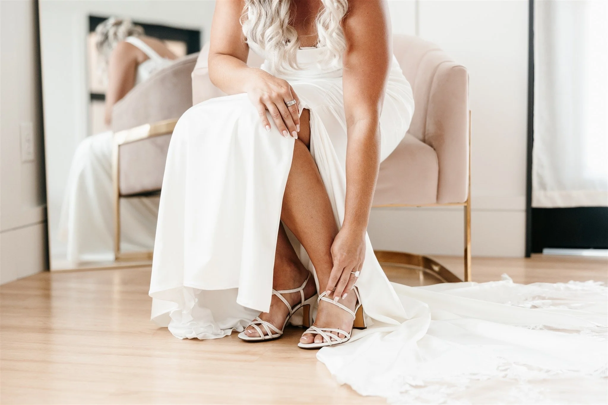 A bride in a white wedding dress is putting on beige strappy high heels while sitting on a pink velvet chair. She is adjusting her shoe and lifting her dress to reveal her leg. The scene is indoors with a mirror reflected in the background.