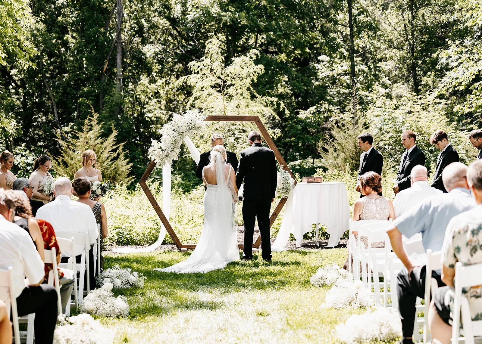 Outdoor wedding ceremony with bride and groom standing under a geometric floral arch, surrounded by seated guests on a grassy lawn with lush green trees in the background.