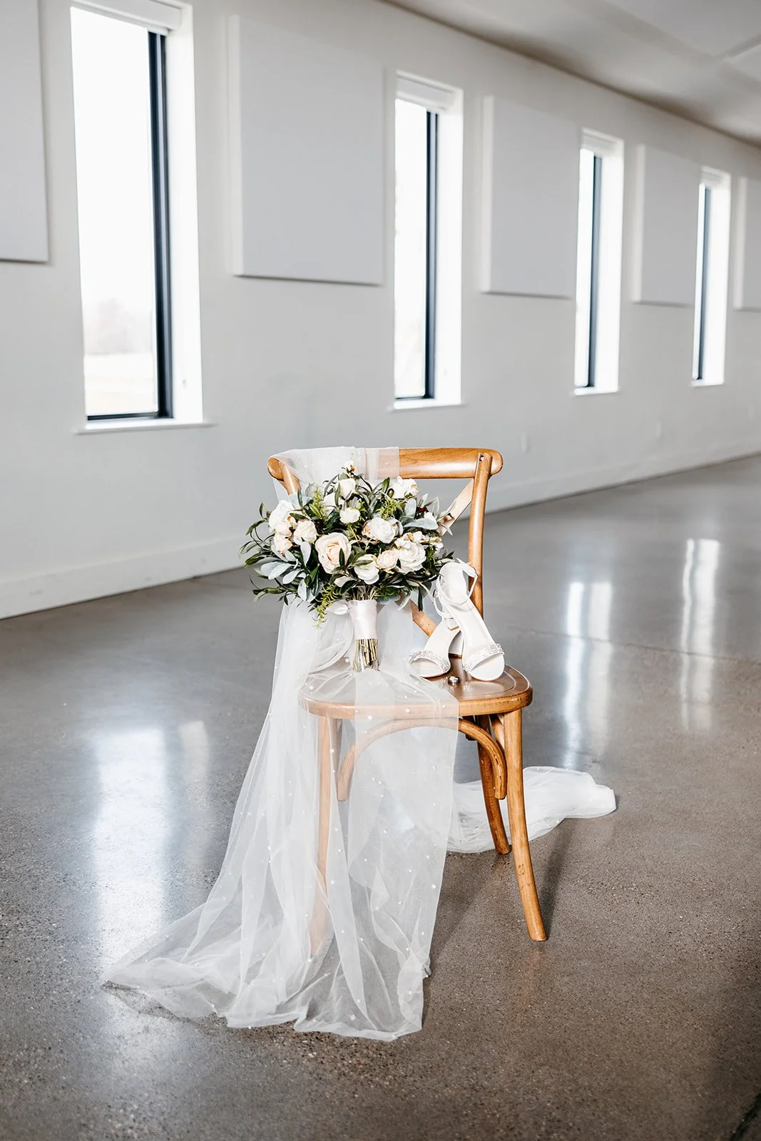A wooden chair decorated for a wedding, holding a bouquet of white flowers, a pair of white high-heeled shoes, and a white veil draped over the backrest; set in a bright, minimalistic room with tall windows.