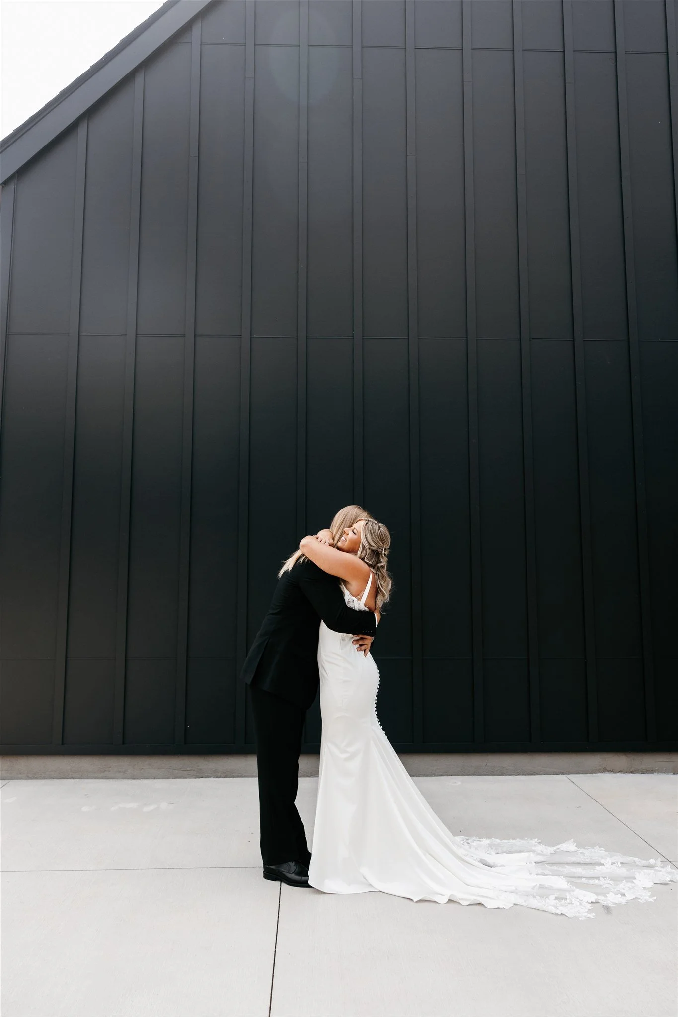 A bride and groom embracing outside against a black building wall, the bride in a white wedding dress with a train and the groom in a black suit.