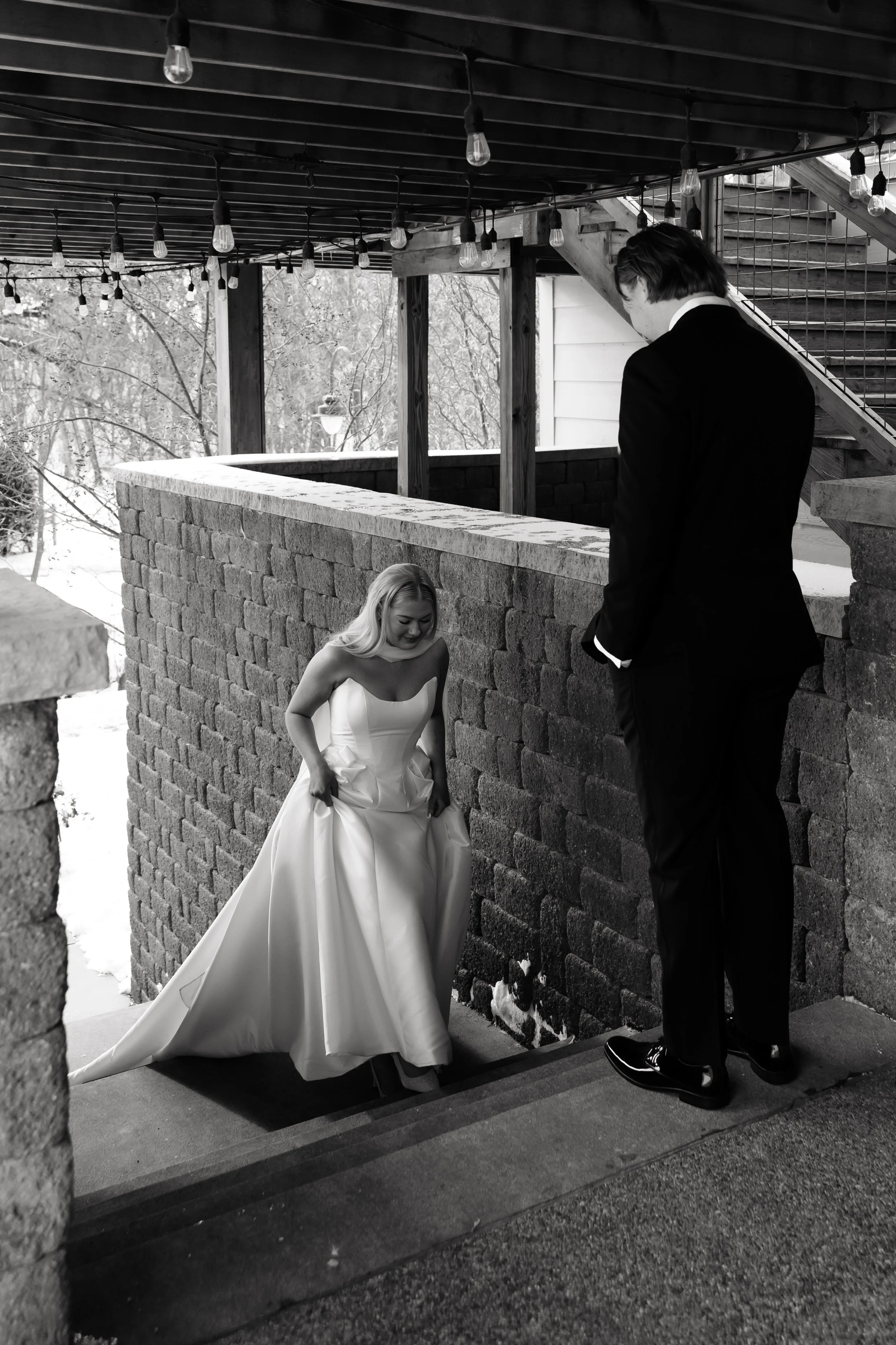 A bride in a strapless wedding gown smiling and holding her dress as she ascends stairs, and a groom in a black suit watching her under a decorated outdoor area with string lights.