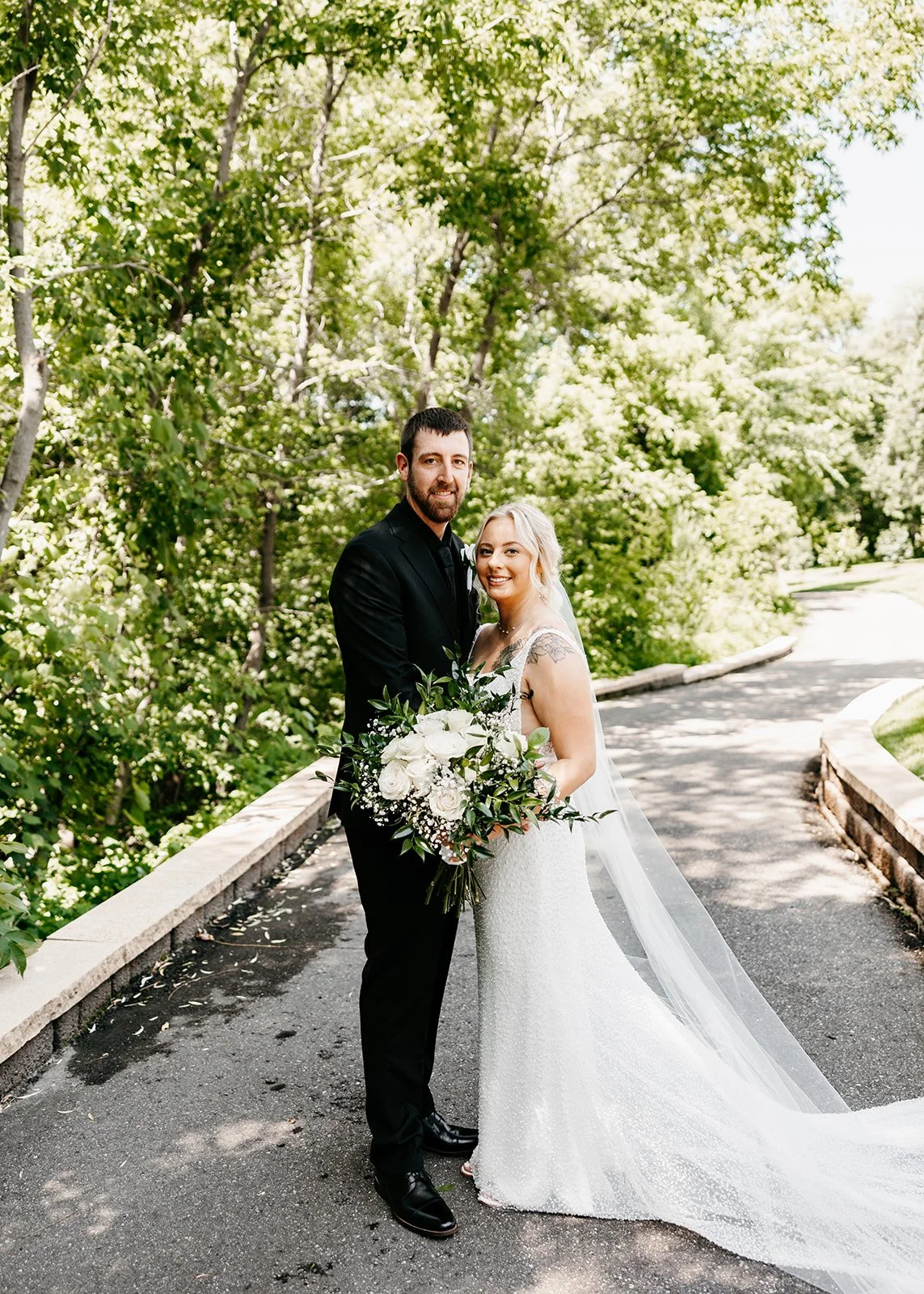 A newlywed couple standing on a paved path surrounded by green trees. The bride is holding a bouquet of white flowers and wearing a white wedding dress with a long veil. The groom is wearing a black suit.