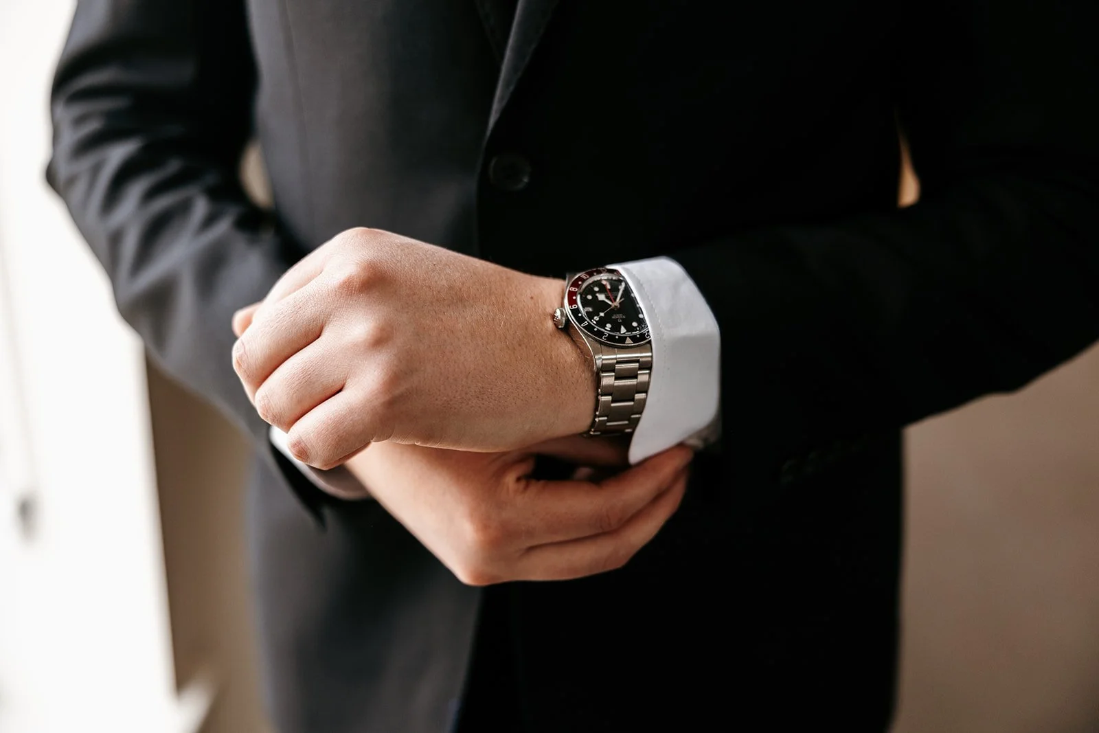 Person adjusting a silver wristwatch with a black dial, wearing a black suit with a white shirt cuff.