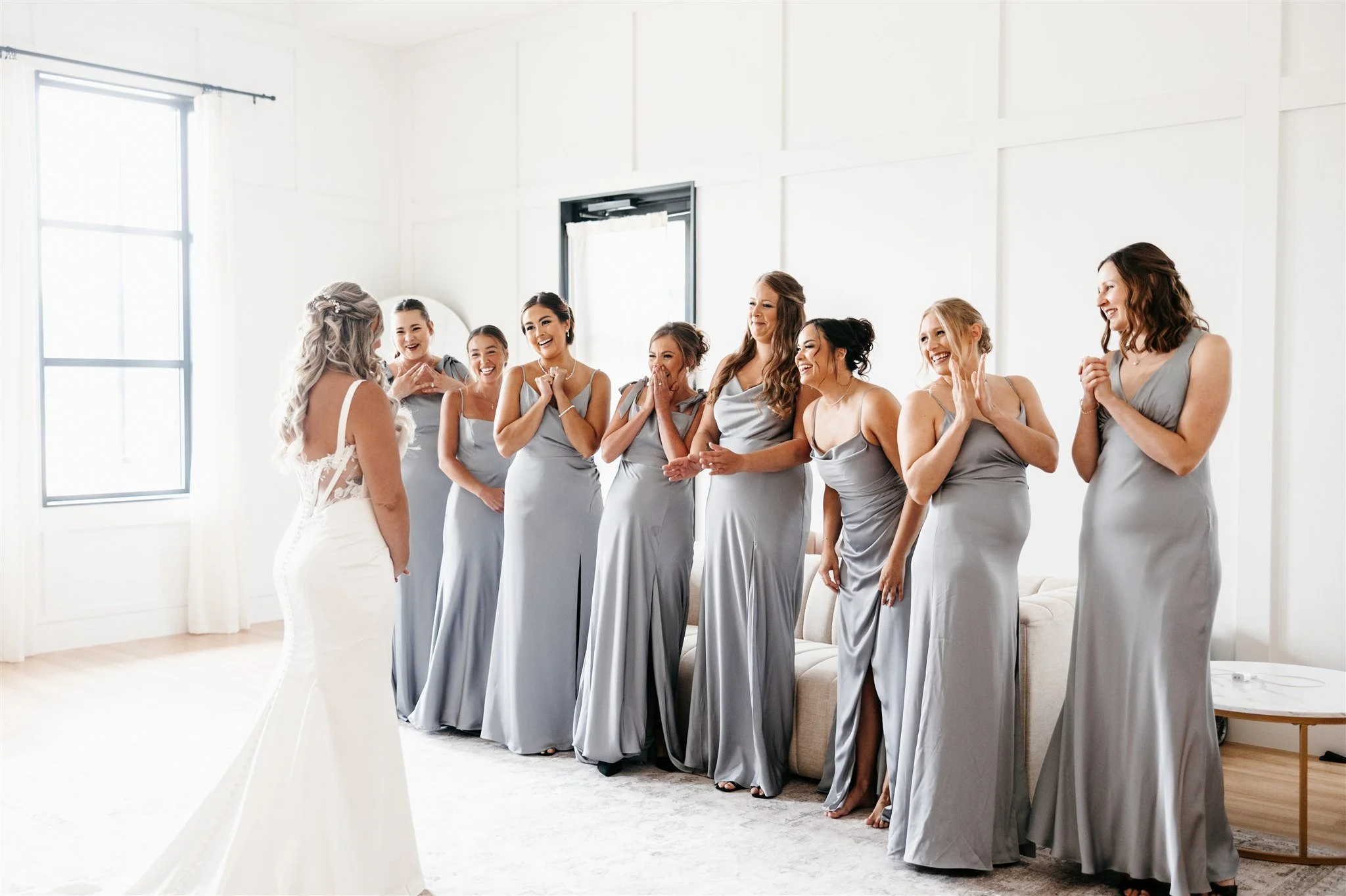 Bridal party in a bright room with white walls and large windows, featuring one woman in a white wedding dress and eight women in matching gray dresses, all smiling and reacting happily.