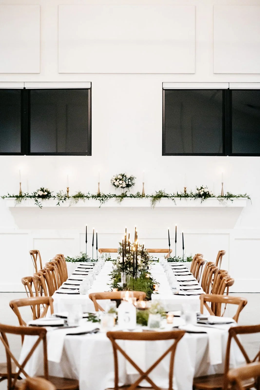Elegant dining table setup with white tablecloths, black candles, and greenery in a modern white room with large black windows.