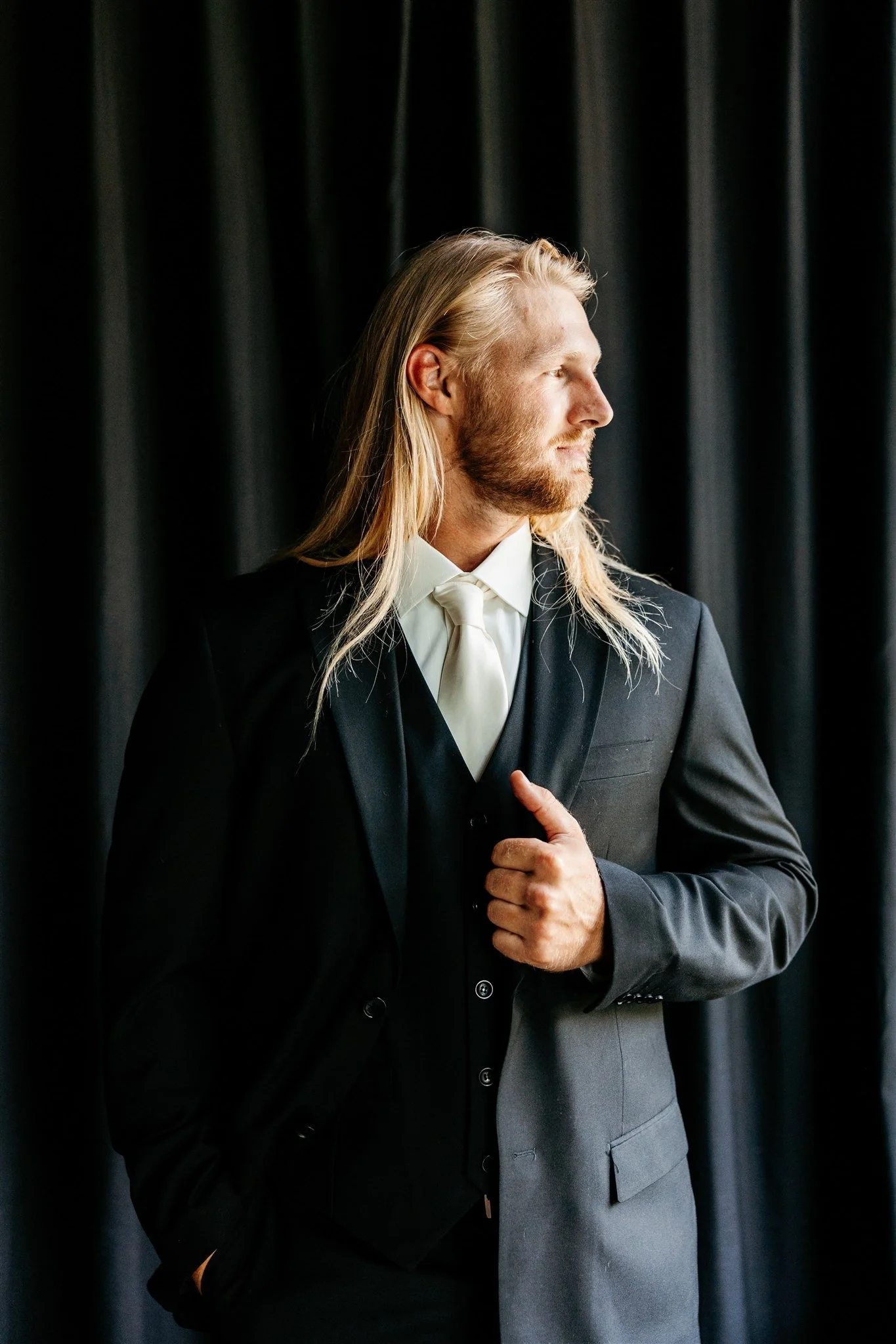 A man with long blond hair and a beard dressed in a black suit and white tie, looking to the side, standing in front of a dark curtain.