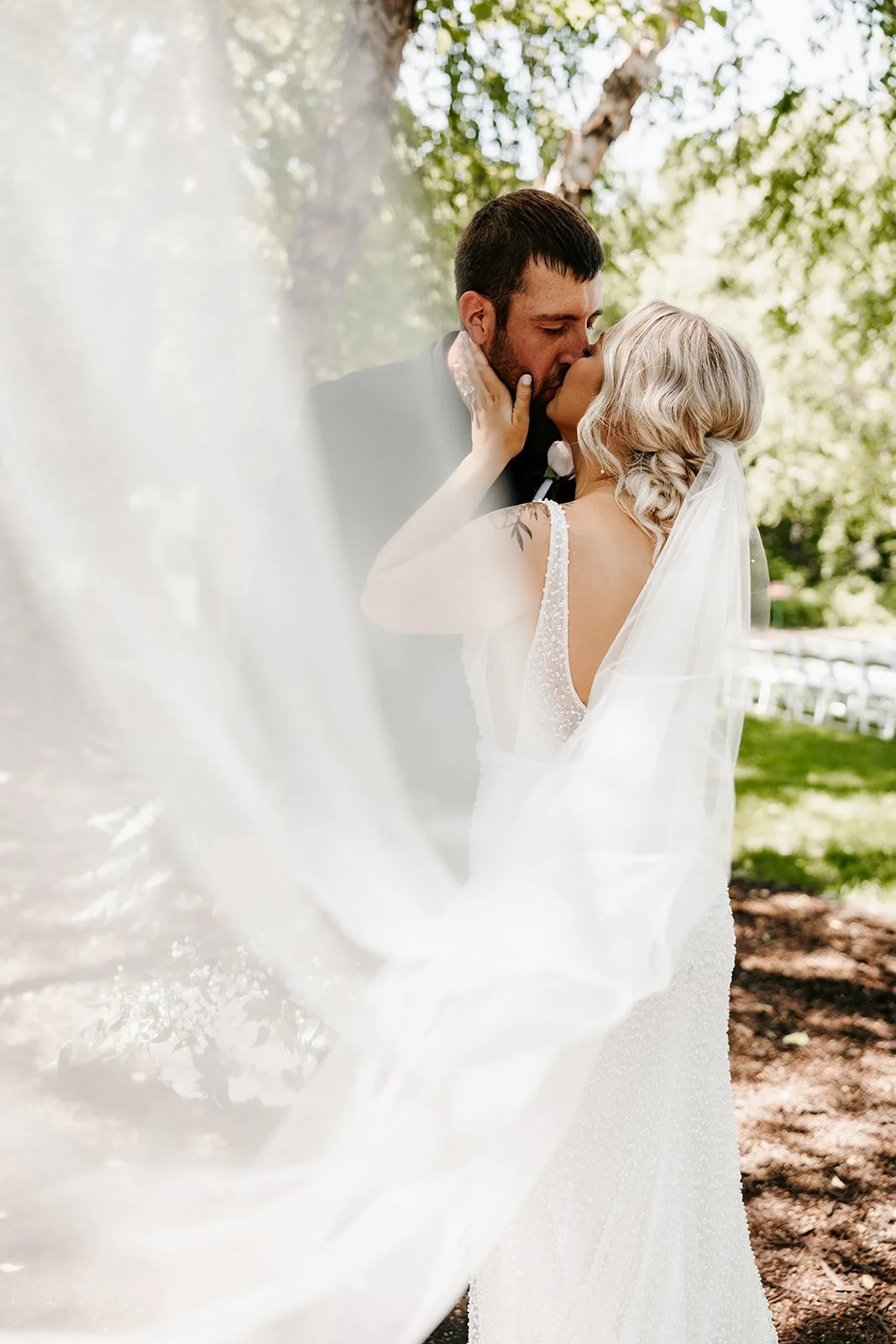 A bride and groom share a kiss outdoors, with the bride's veil flowing in the wind, surrounded by greenery.