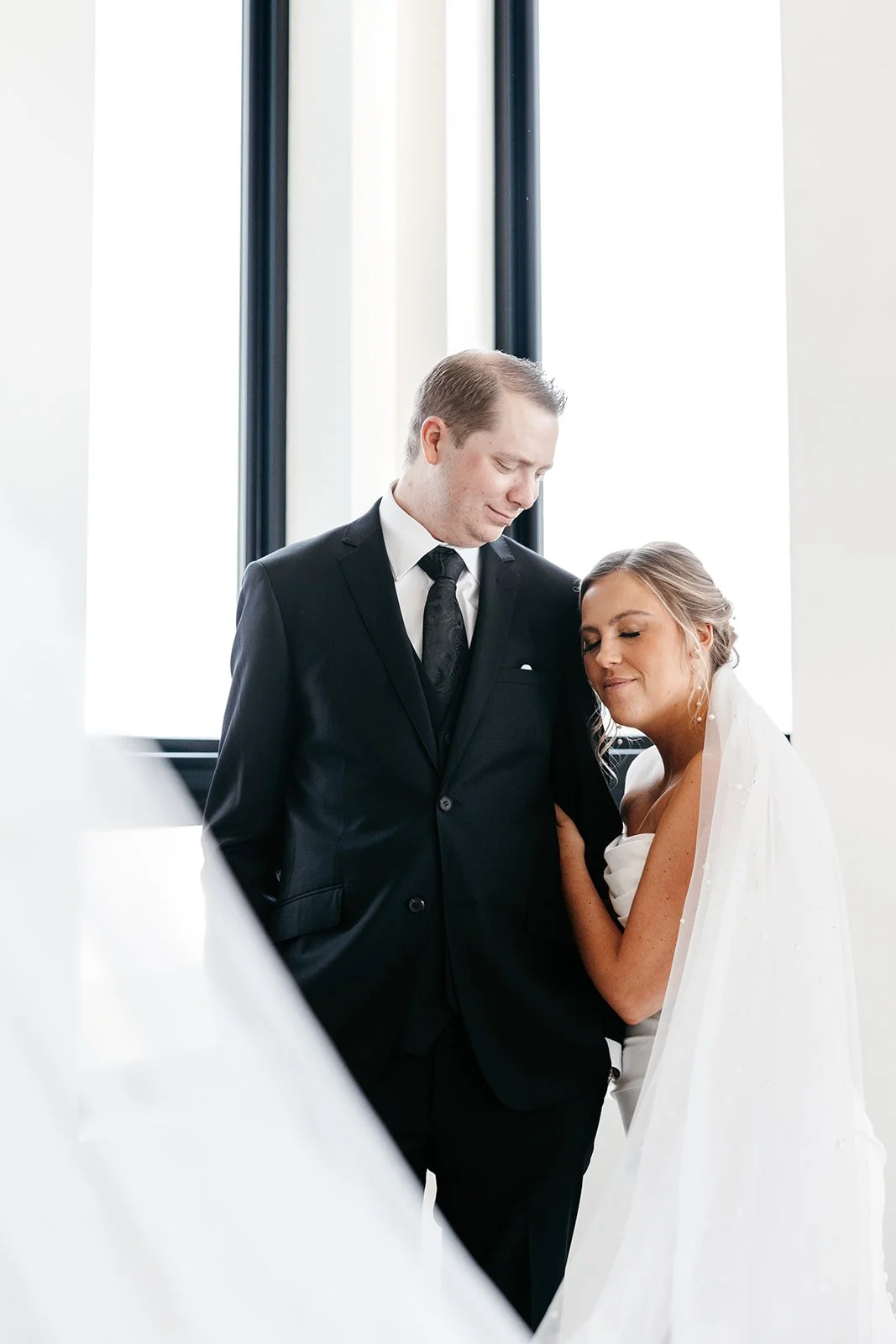 Bride and groom share a tender moment indoors, with the bride resting her head against the groom's chest and eyes closed.