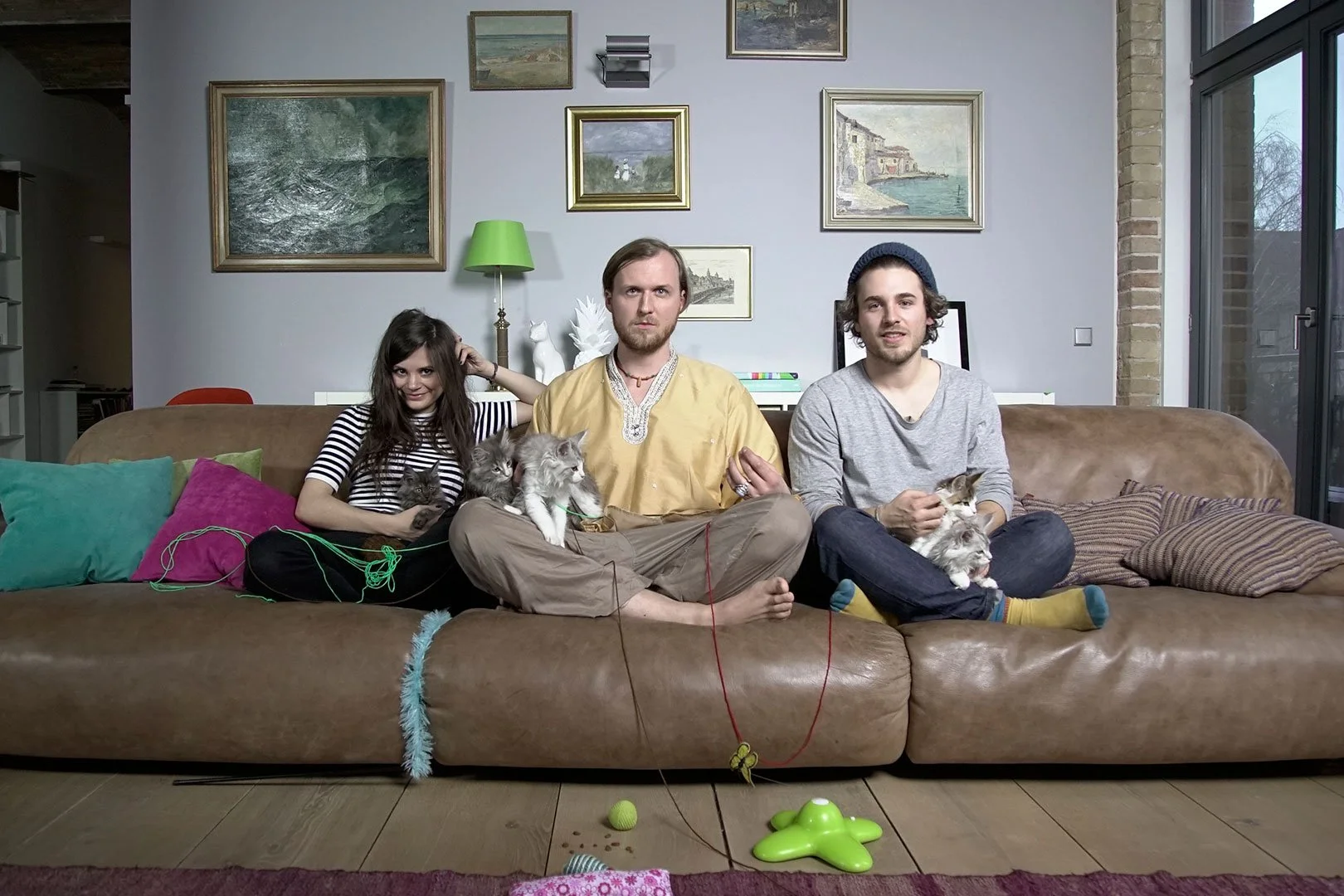 Drei Menschen sitzen auf einem braunen Sofa, umgeben von Katzen und Spielzeugen, in einem Wohnzimmer mit Bildern an der Wand und großen Fenstern.