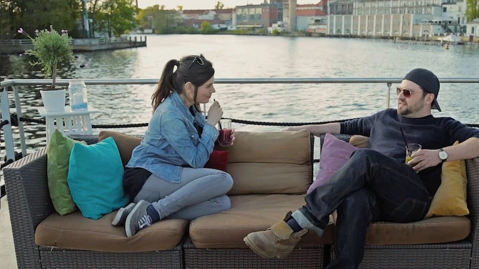 Zwei Menschen sitzen auf einem Sofa auf einer Terrasse mit Blick auf den Fluss, trinken Getränke und unterhalten sich.