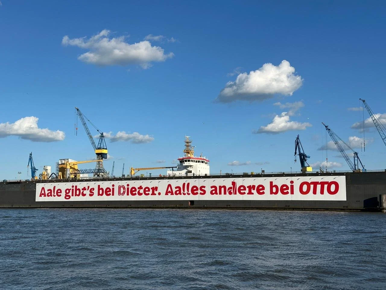 Großes Schiff im Hafen mit einem weißen Banner, auf dem auf Deutsch steht: 'Aale gibt's bei Dieter. Alles andere bei Otto'.