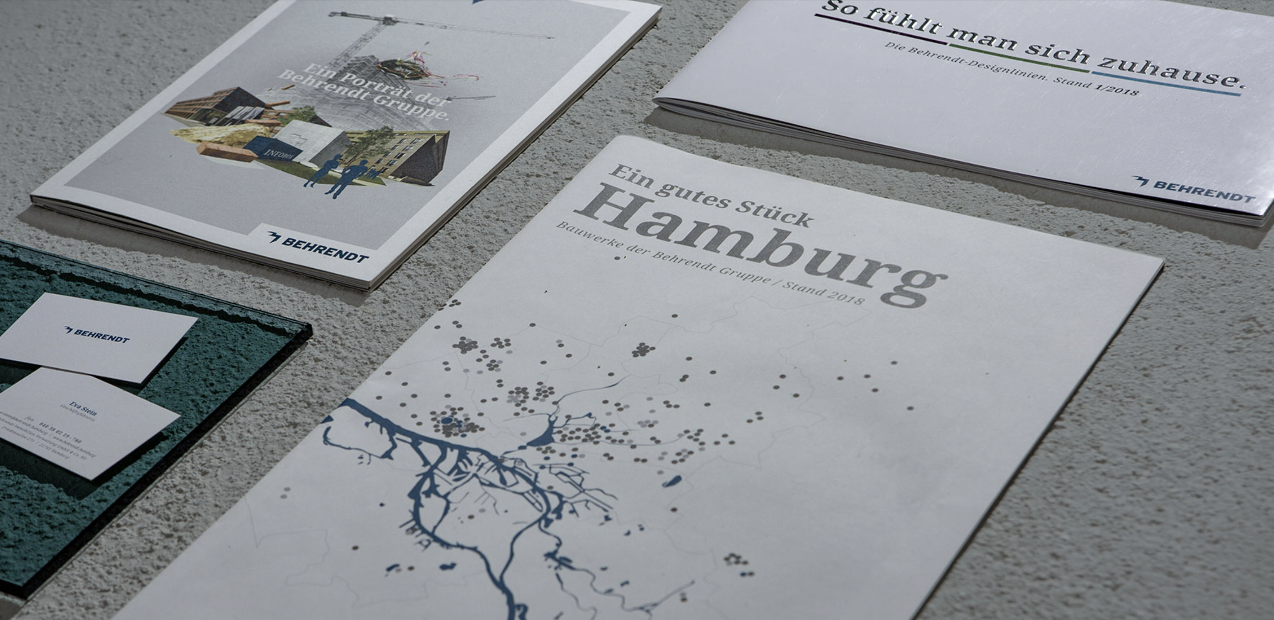 Drucke und Broschüren der Behrendt-Gruppe, darunter eine Karte von Hamburg, eine Broschüre mit einem Stadtmodell, und Visitenkarten, auf einer Steinoberfläche.