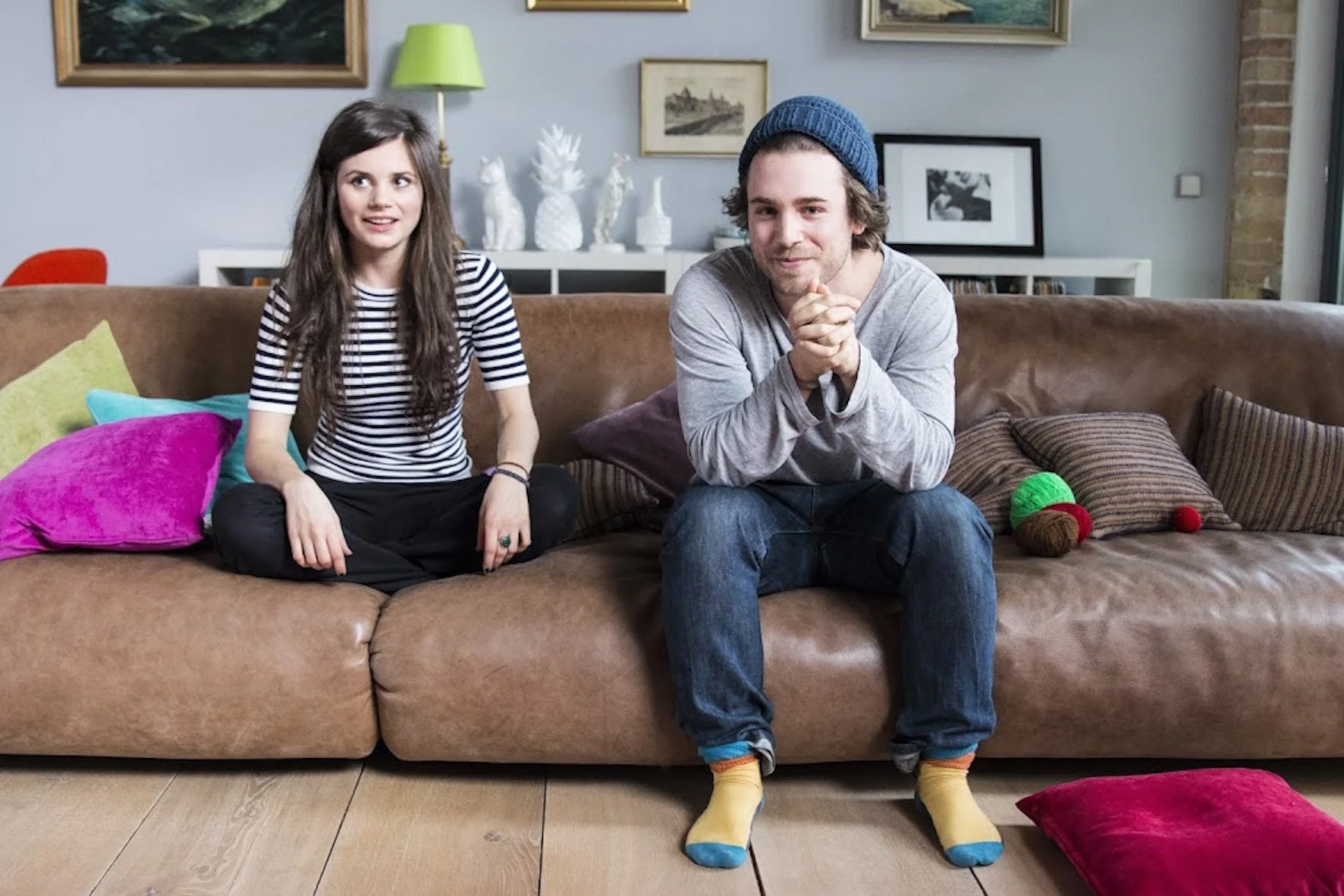 Ein junger Mann und eine junge Frau sitzen auf einem braunen Sofa in einem Wohnzimmer, umgeben von bunten Kissen, mit gestreiften Hemden, einer Mütze und Socken, und wirken entspannt.