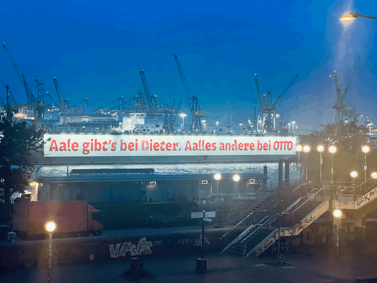 Nachtaufnahme eines Hafens mit Kränen im Hintergrund. Es ist eine große Werbetafel mit rotem Text auf einer Brücke, die ungefähr sagt: "Aale gibt's bei Dieter. Alles andere bei OTTO".