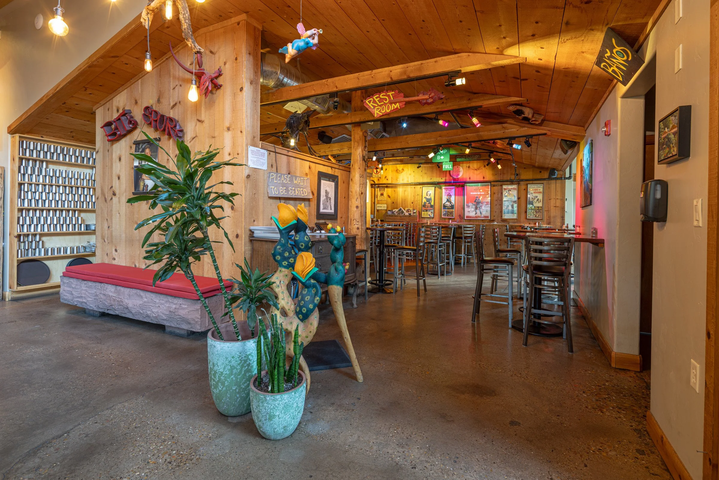Interior of a cozy bar or restaurant with wooden walls, bar stools, framed posters, and potted plants. Decorations include hanging lights, a sign that says 'Please wait to be seated,' and other colorful signs.