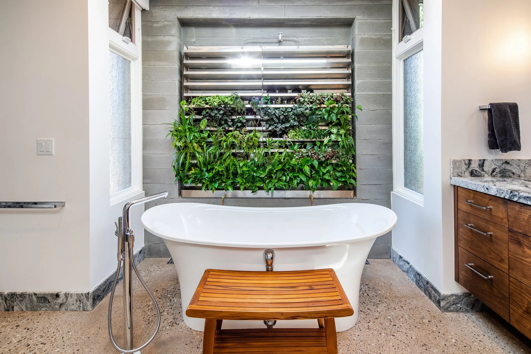 Bathroom with a bathtub positioned in front of a vertical garden on a gray wooden wall, flanked by two windows, a granite countertop with drawers on the right, and a towel hanging on the wall.