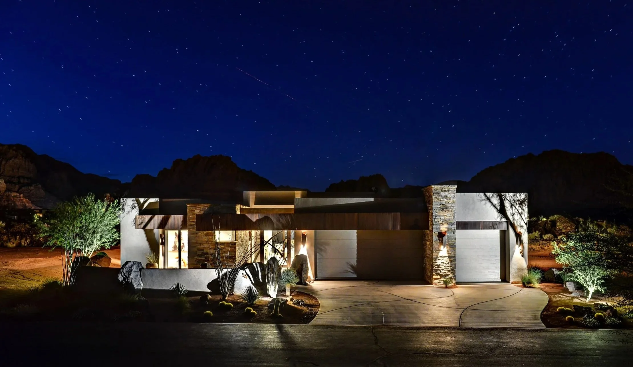 Nighttime view of a modern house with outdoor lighting, surrounded by desert plants and rocks, under a starry sky.