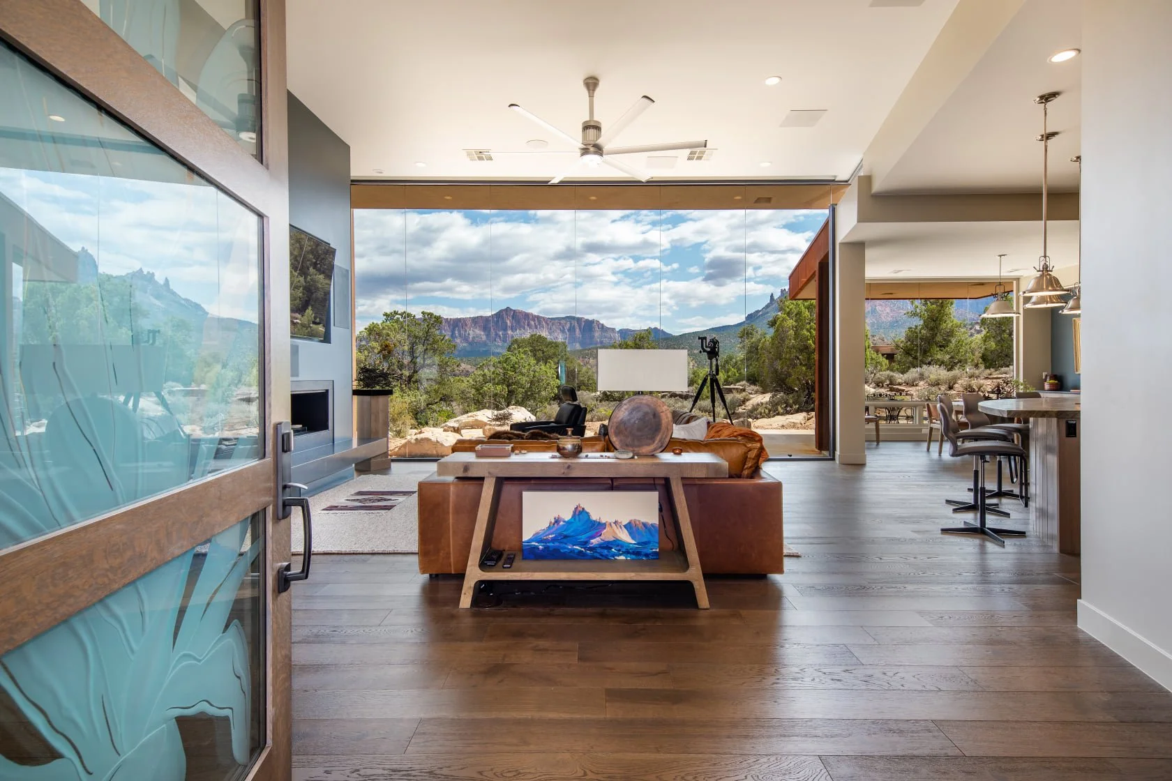 Open-concept living room with large glass windows showcasing mountain and desert landscape, wooden floors, modern furniture, a ceiling fan, and mounted TV