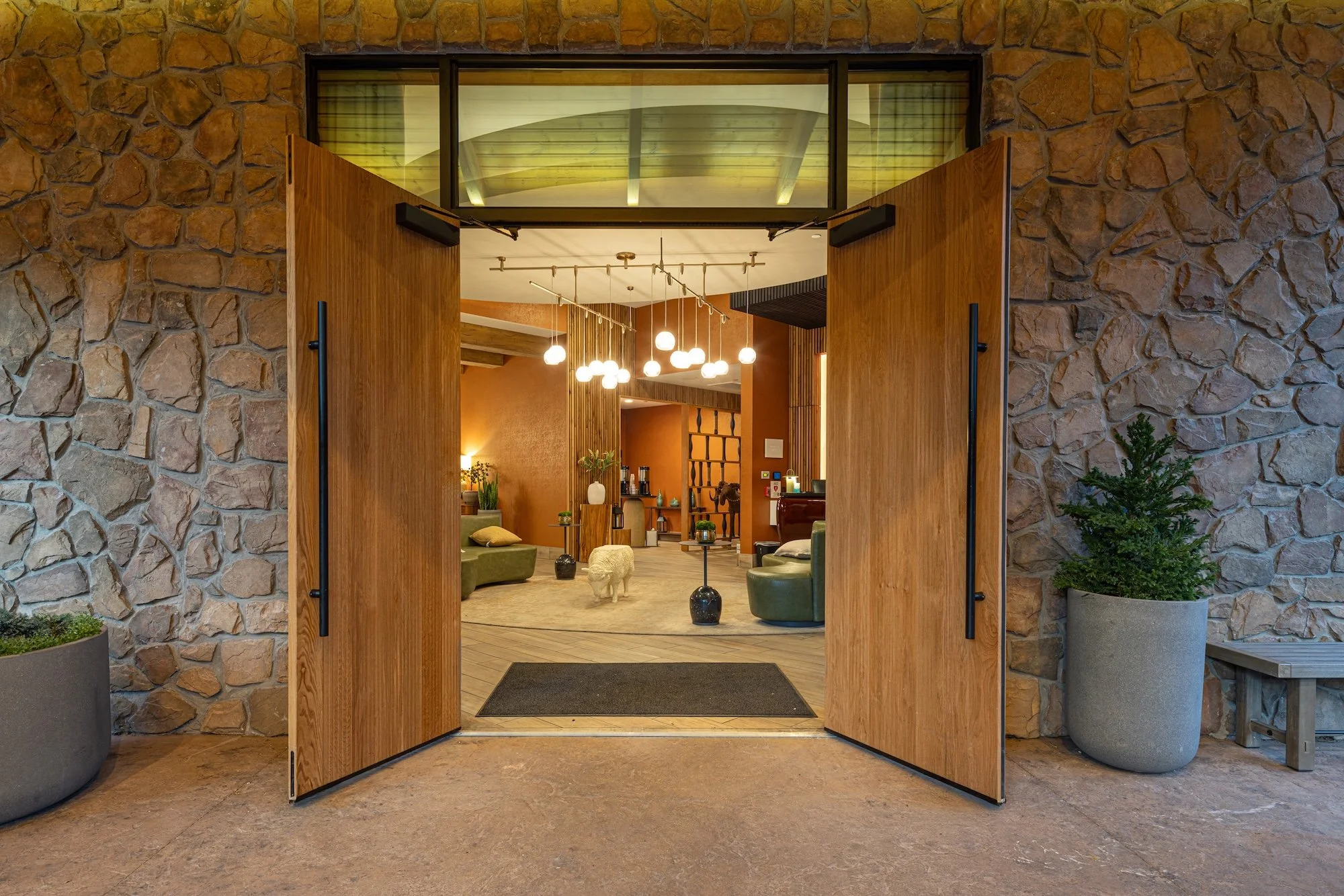 Open wooden doors leading to a warmly lit interior space with modern decor, hanging pendant lights, plush seating, and decorative plants.