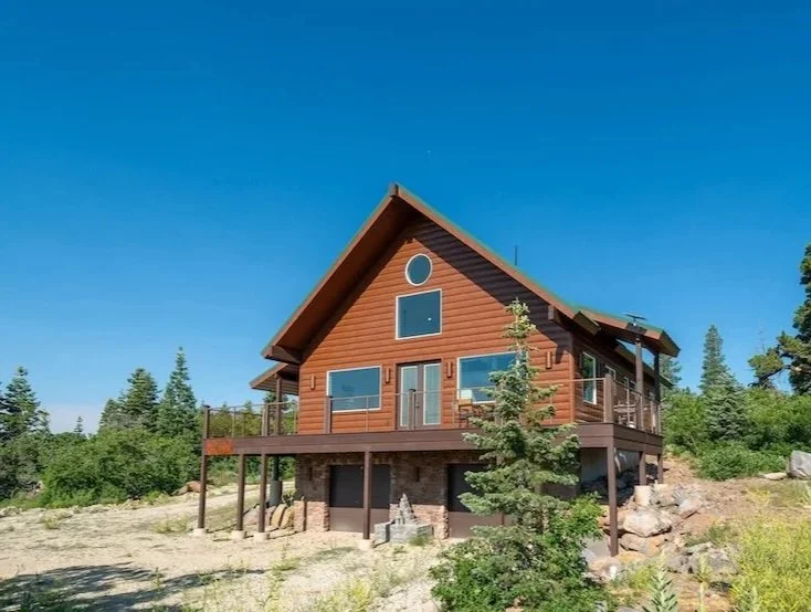 A wooden house built on stilts with a deck, set against a backdrop of trees and a clear blue sky.