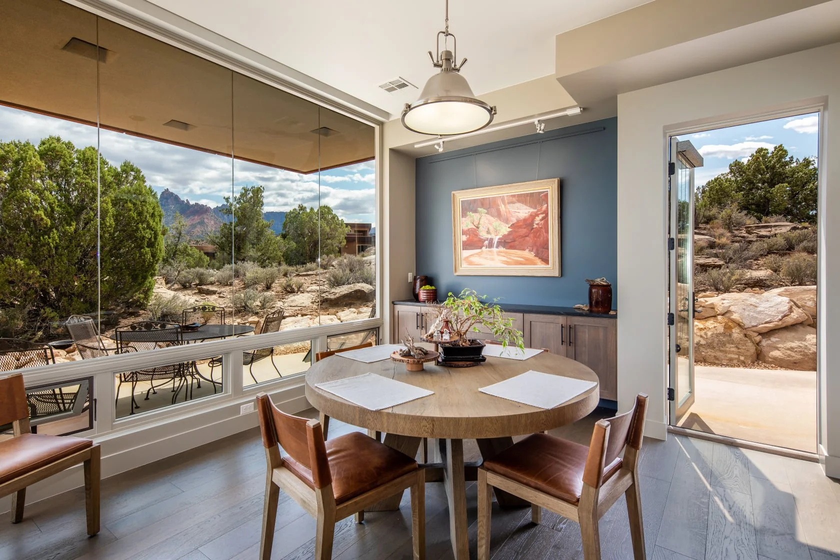 Dining room with a wooden round table and four chairs, facing large windows showing a desert landscape with trees, rocks, and mountains, and a door leading outside.