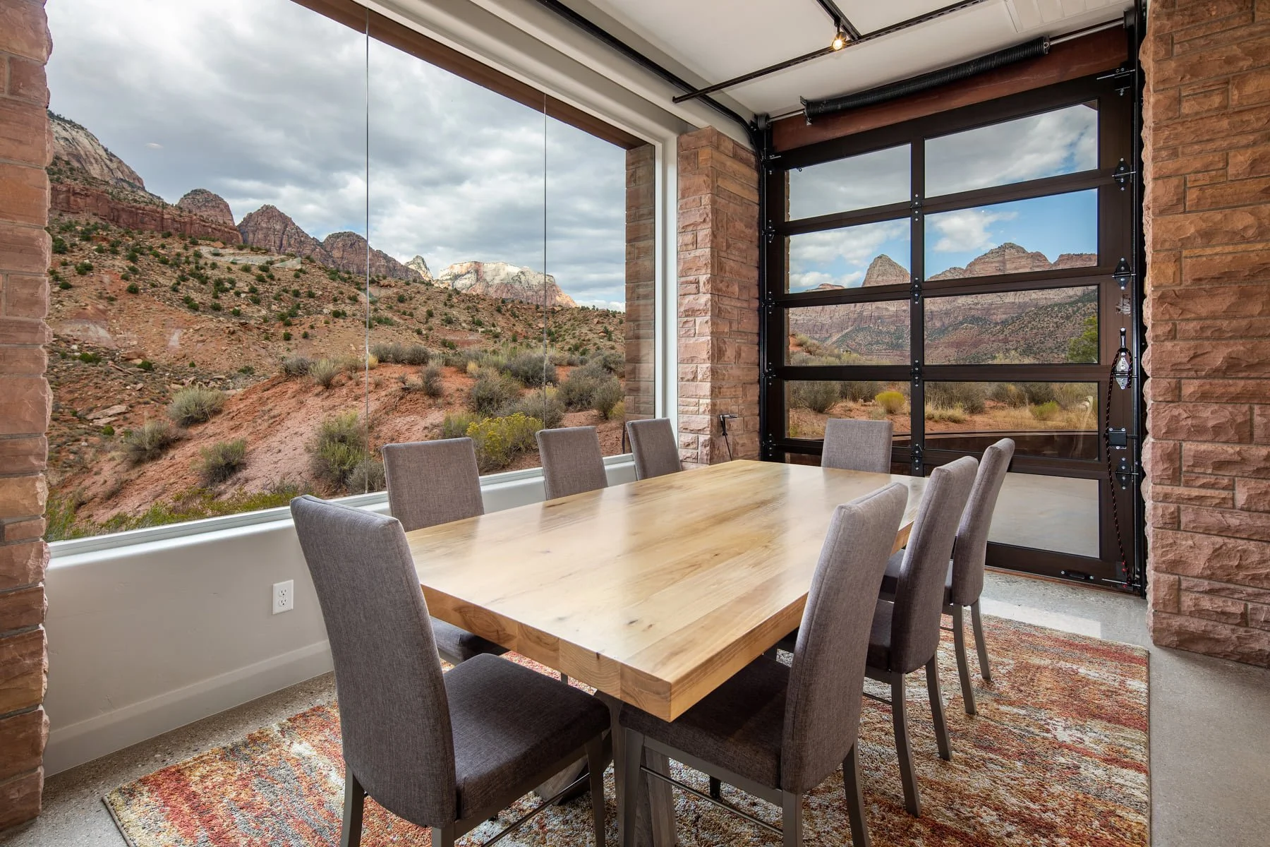 Modern dining room with large window and view of desert mountains, featuring a wooden dining table, eight upholstered chairs, exposed brick wall, and an industrial-style garage door.