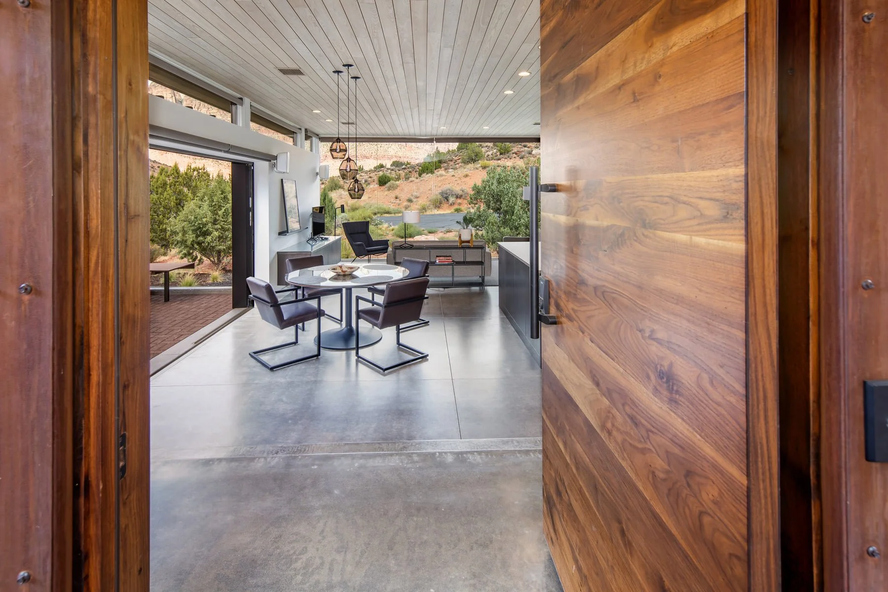 Open doorway leads into a modern living room with large windows overlooking a hilly landscape with trees. The room features a round dining table with four chairs, a black armchair, a sofa with a side table, and pendant lights hanging from a wooden ceiling.