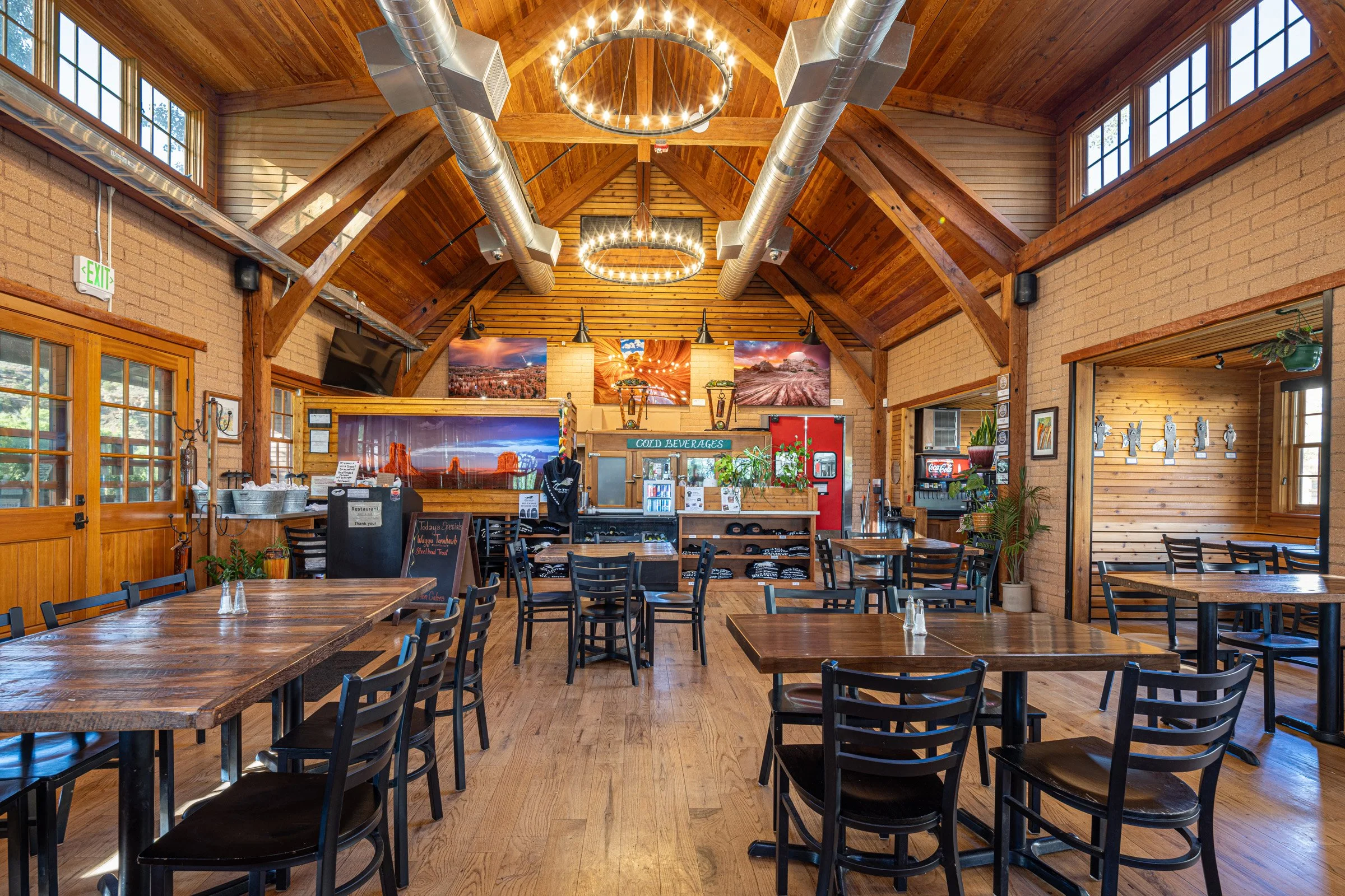 Interior of a cozy restaurant with wooden walls, ceiling beams, and multiple television screens on the walls. There are several empty wooden tables and chairs, a small serving area, and decorative lighting fixtures hanging from the ceiling.