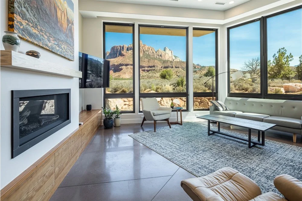 Living room with large windows showing a desert landscape with rocky formations, a cream sofa, an armchair, a modern coffee table, a large flat-screen TV, a fireplace, and decorative items.