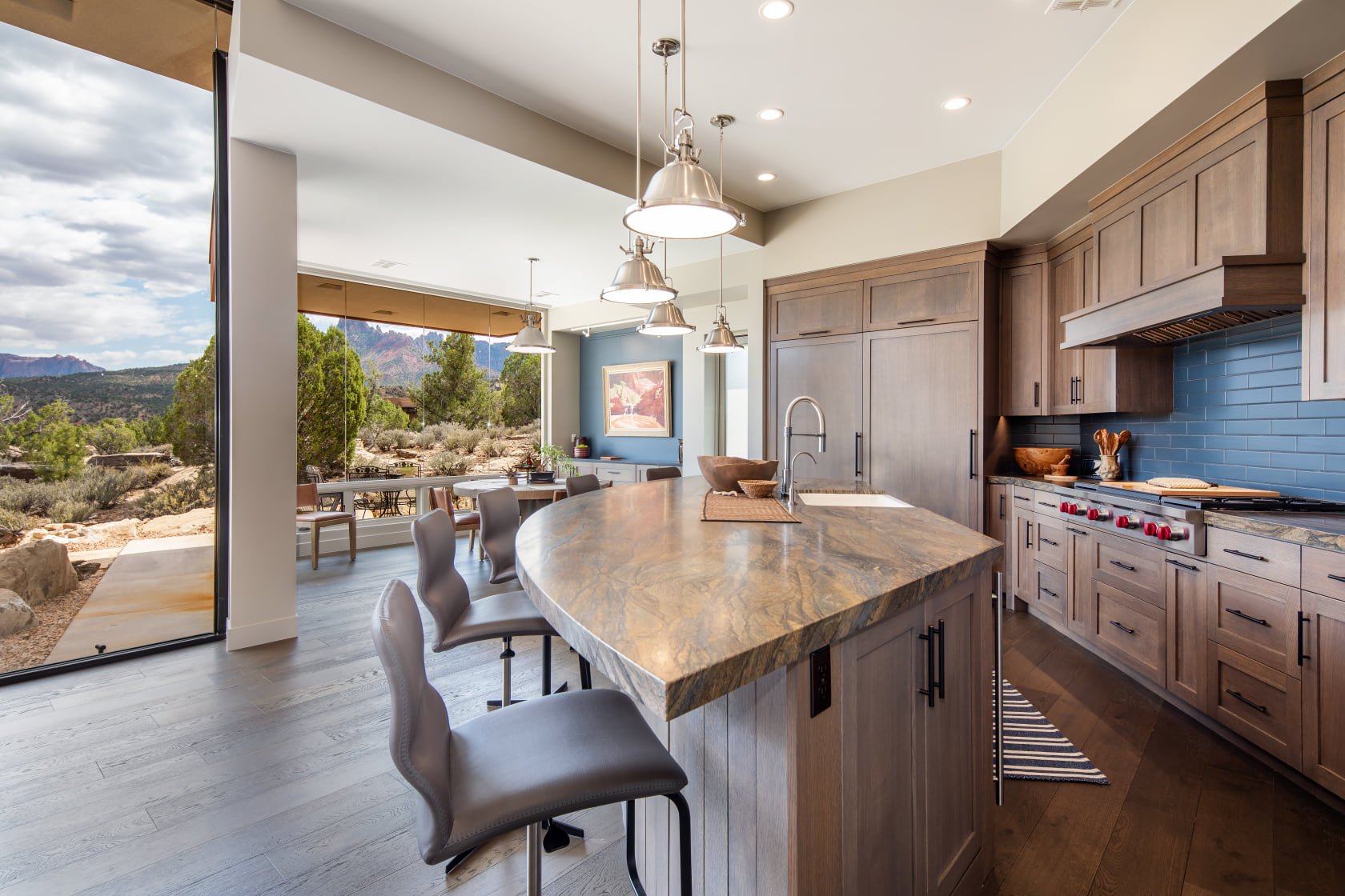 Modern kitchen with large island, wooden cabinets, blue tiled backsplash, stainless steel stove, and glass sliding doors opening to mountain view.