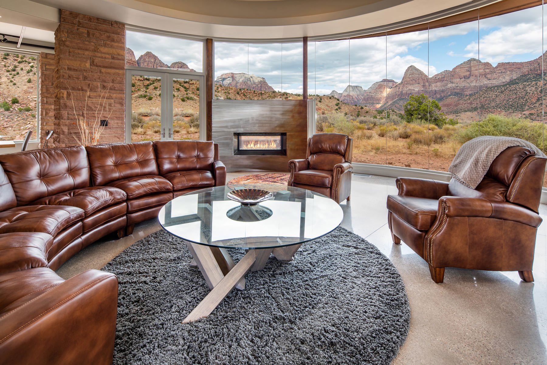 Living room with large windows overlooking desert landscape and mountains, featuring brown leather sectional sofa, matching armchairs, glass coffee table, stone fireplace, and plush gray rug.