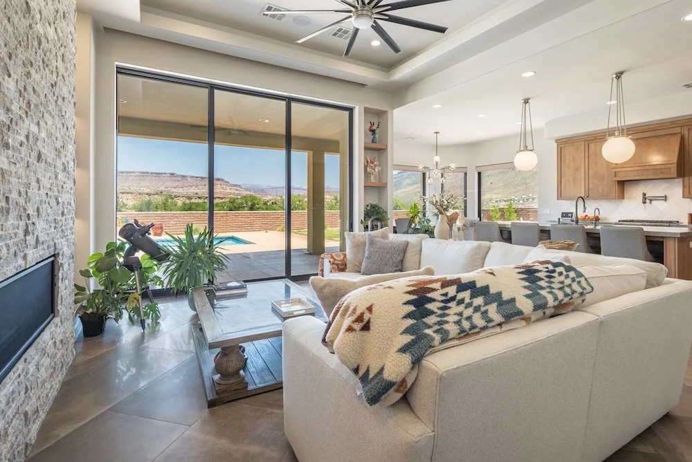 Living room with large glass sliding doors leading to outdoor patio and swimming pool, stone accent wall, beige sectional sofa with blanket, indoor plants, modern chandelier, open kitchen with wooden cabinets and bar seating, mountain view through windows
